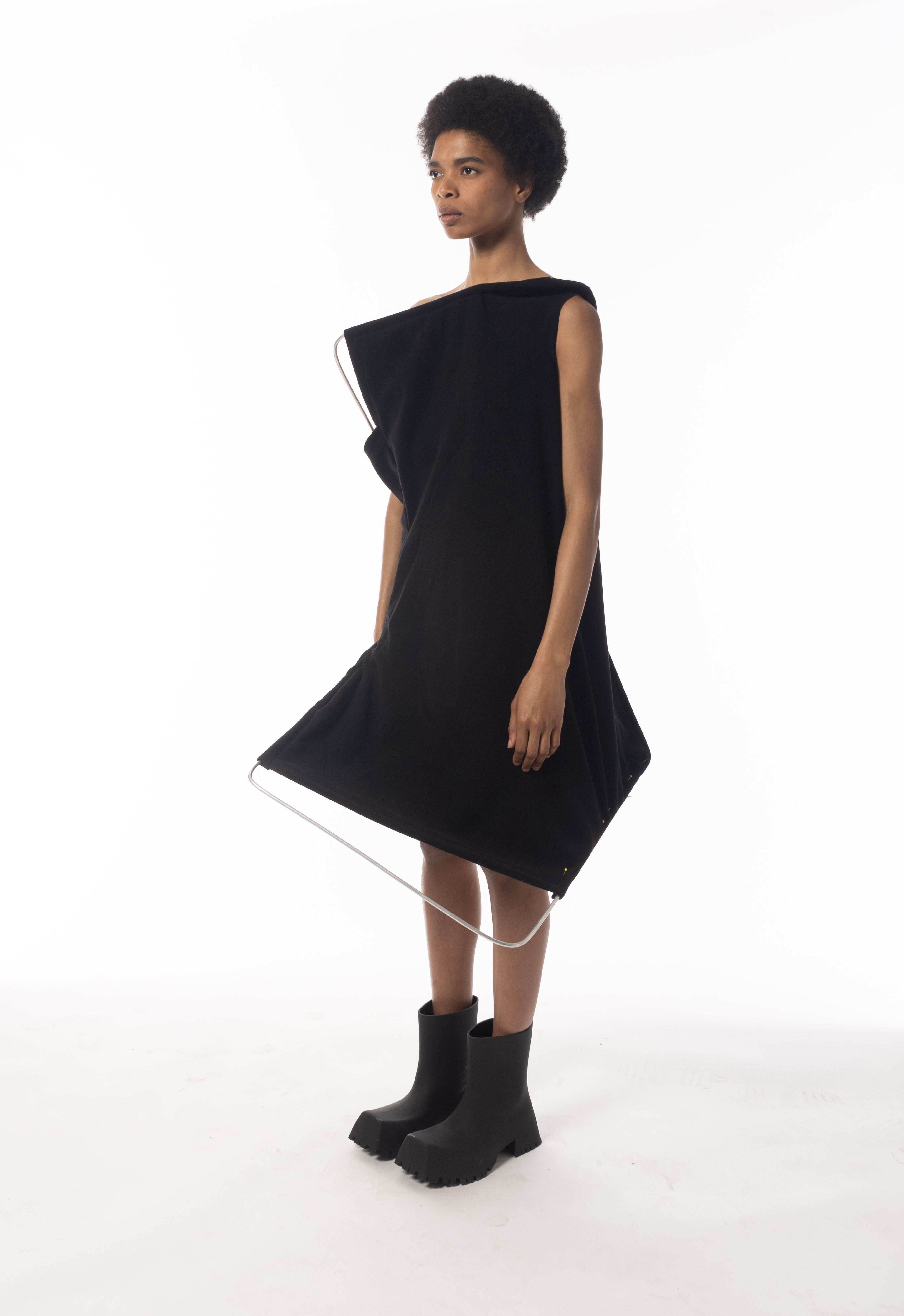 Front view of a woman wearing a Mid-century interior-inspired, black foldable knee-length wool dress with a metal frame stretching the structure. The model is wearing black boots. The background is white.