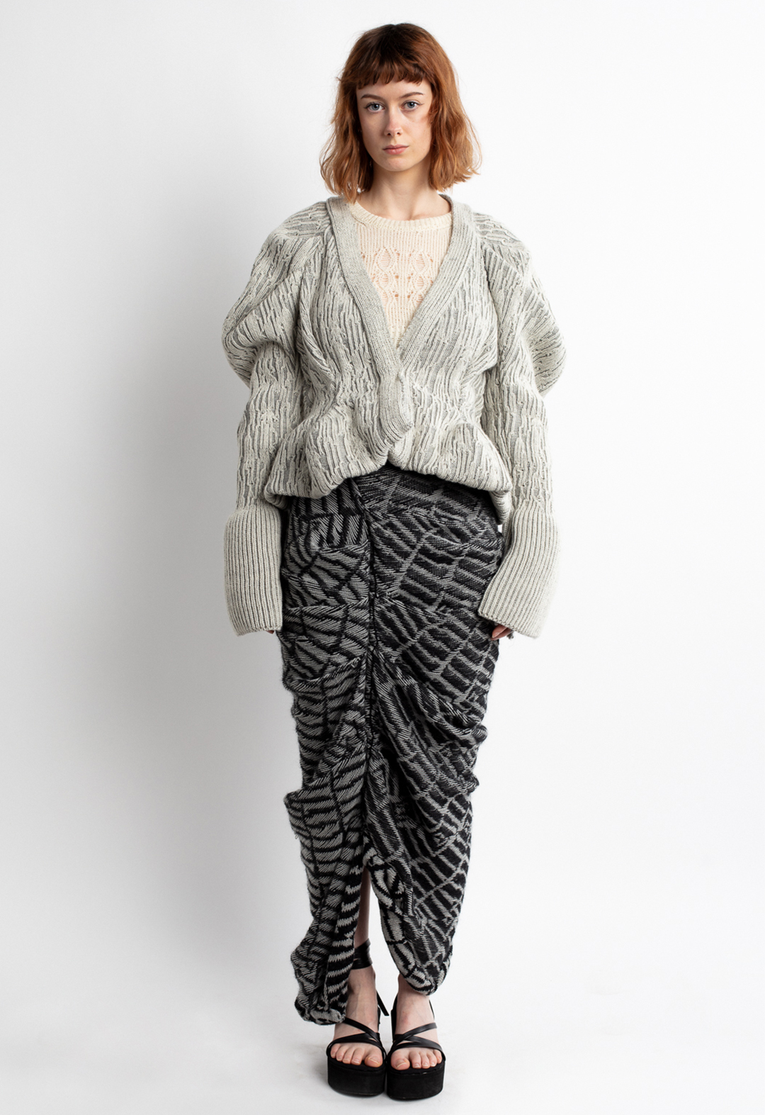 This look is inspired by many aspects of old and dried trees, such as bark textures, twisted roots, and shapes of branches. This jacket's shape is three-dimensional, with its draped sleeves and waist to hemline. The bottoms of the sleeves are knitted with full-cardigan fabric. The cropped inner top is fitted to her bodice, and the high-waisted skirt is draped asymmetrically. The designed ribbed fabric of the jacket is knitted on Stoll. The sweater is knitted on every other needle, and the skirt is knitted as jacquard fabric with a designed punch card on a Brother knitting machine. For the jacquard fabric of the skirt, the wrong side is used to show its floated parts. 