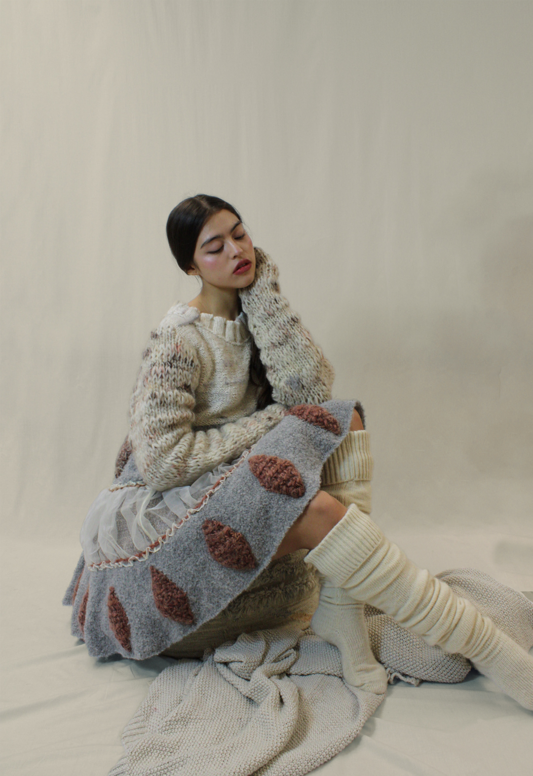 Photo of a girl wearing a multitextural earth-toned babydoll dress with knee high socks. She is sitting on a bean bag and appears to be lost in thought. The background is a warm white.