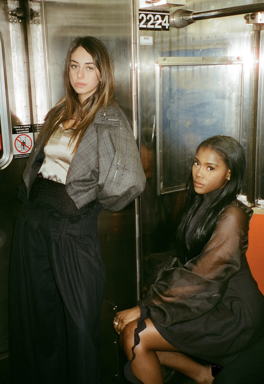The photo shows two models on a subway. The model standing is wearing a wool plaid blazer with a silk camisole and wool pants. The model sitting beside her is in a dress with organza sleeves.