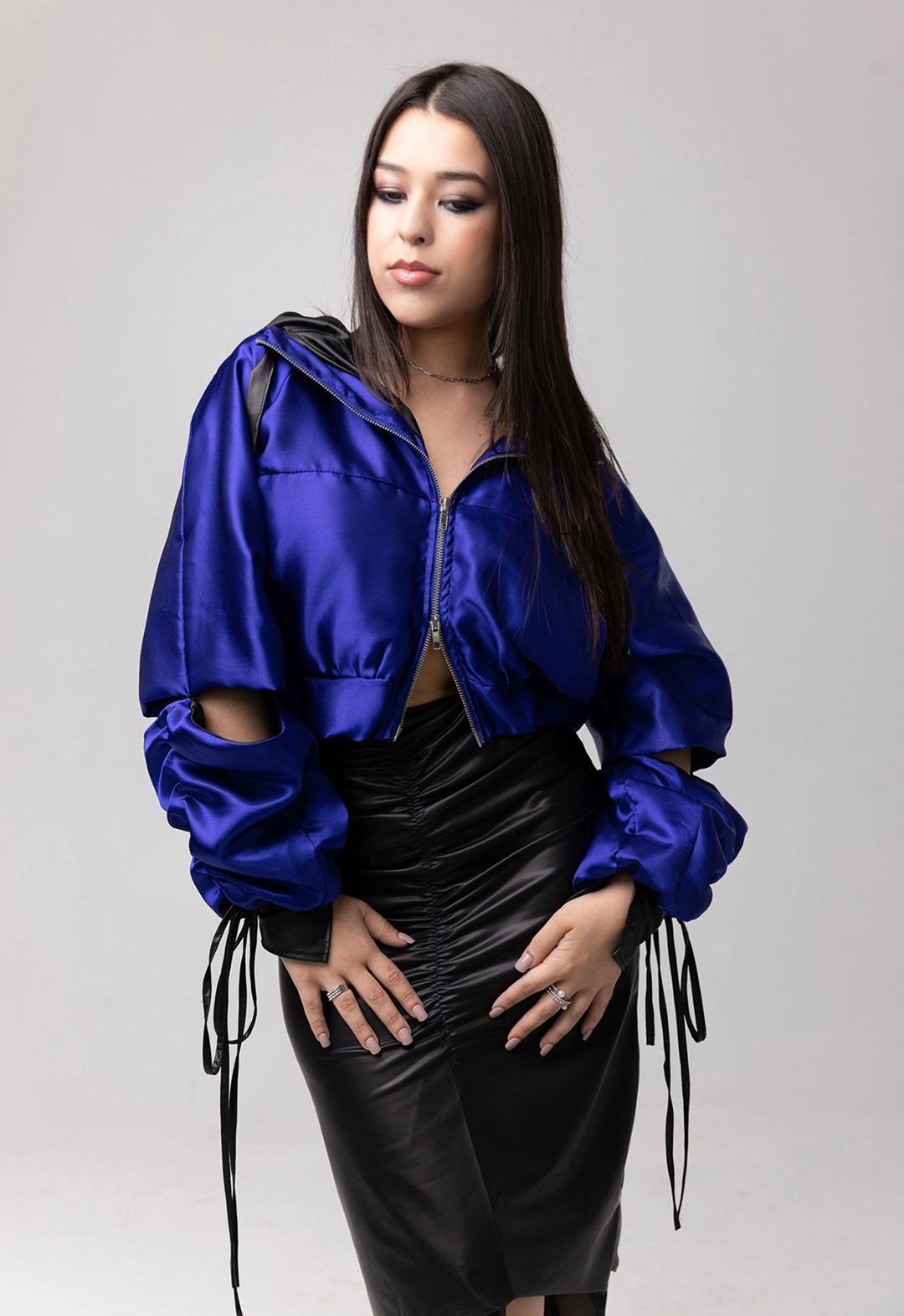 This is the front view of a model wearing a royal blue jacket and a black skirt with gathers on center front. The jacket has cut-outs on the elbow and drawstrings hanging from the cuff. The model has both her hands on her thighs. The background is gray. 