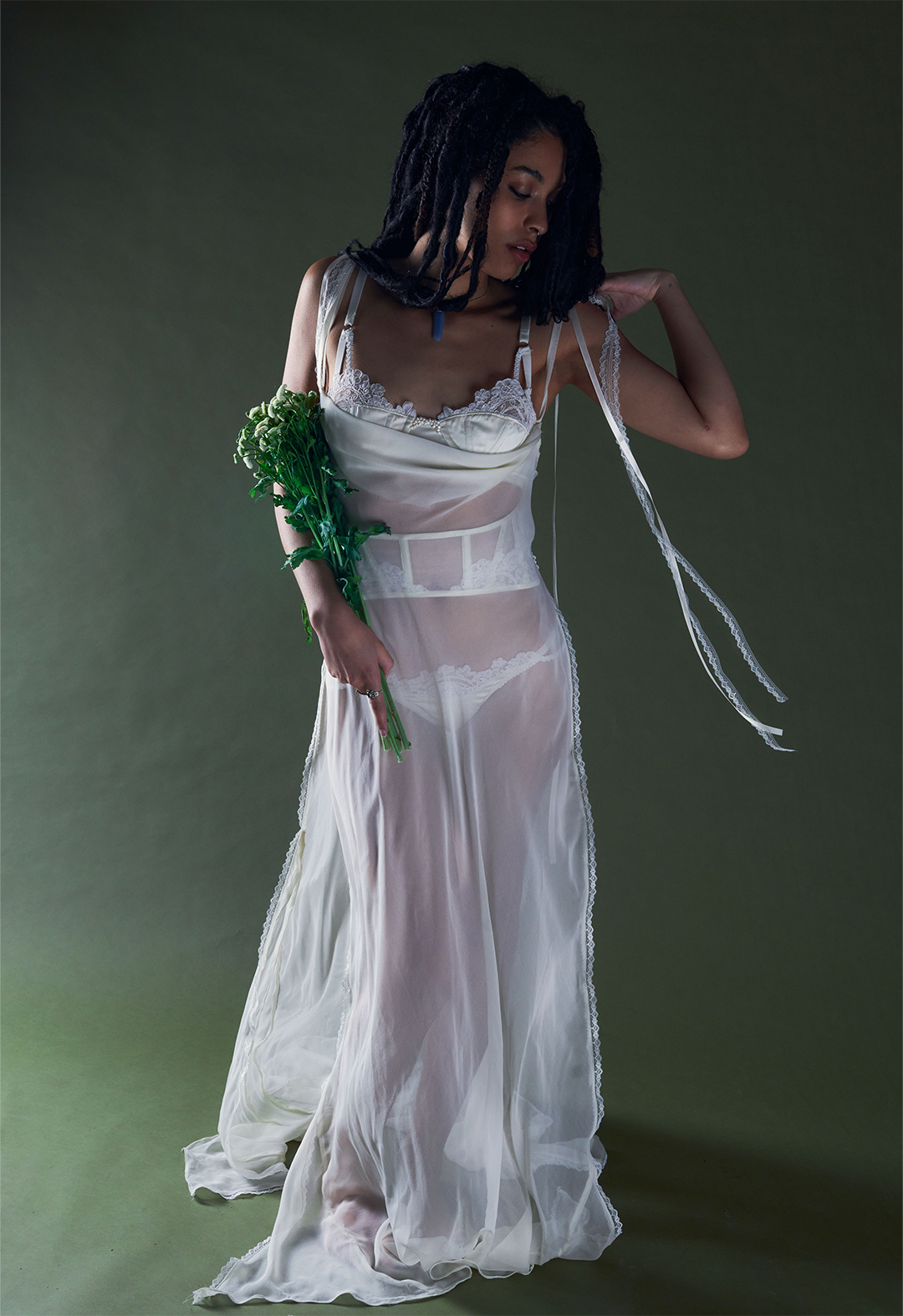 This image features a model in a long pearl-white bias gown with a cowl neck, open side seams and lace detailing. It is paired with a bra and panty set and matching waist corset, embellished with lace and pearls. The image is set in a green background.