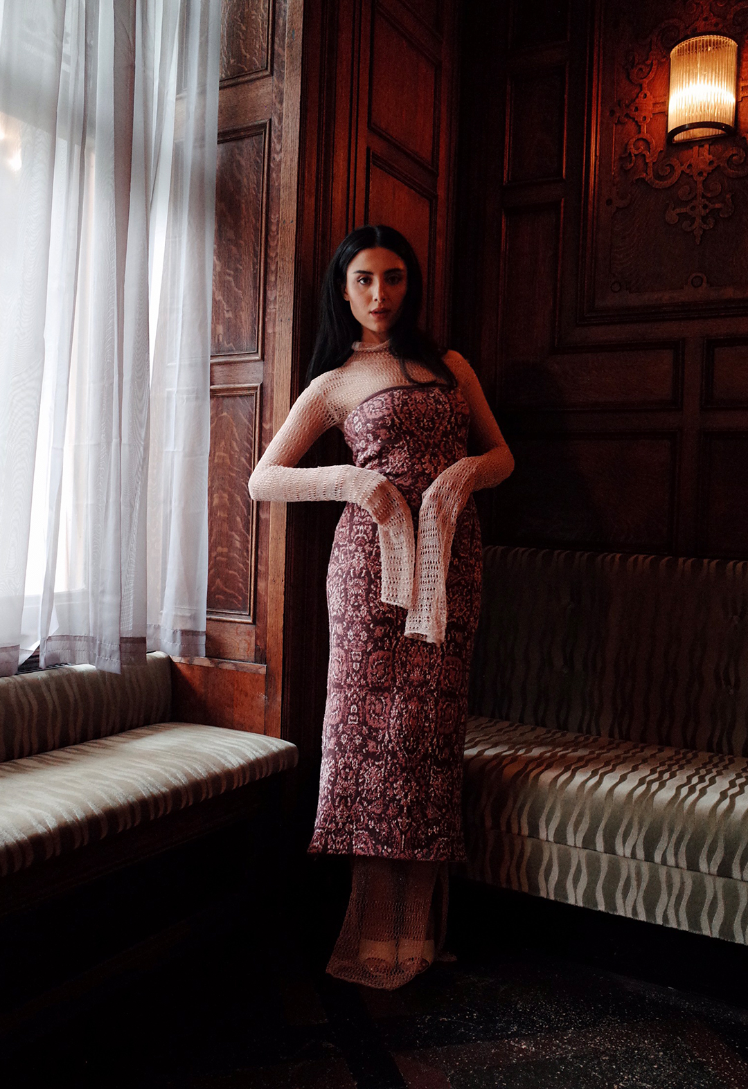Photo of a model wearing a carpet inspired tube dress layered over sheer pointelle dress. She is standing near a window with a sheer curtain over it.