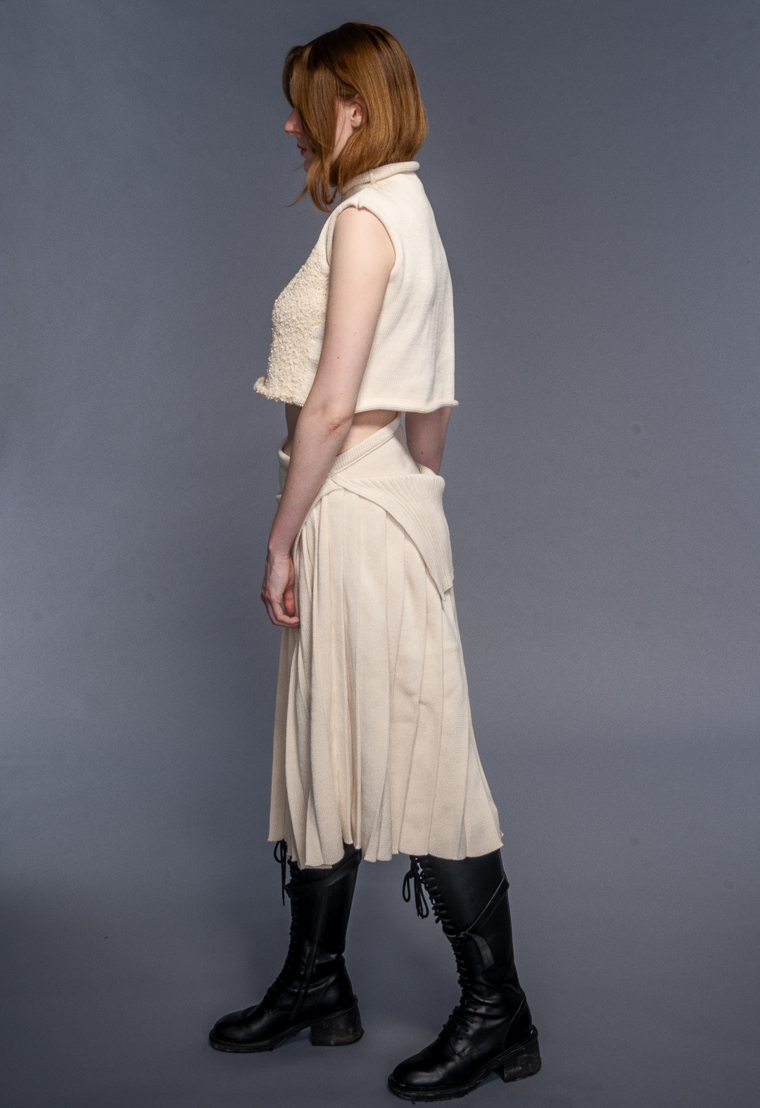 Model wears funnel neck top with misshaped intarsia and irregular pleated skirt in organic cotton. Her hair is tucked in the sweater and she is looking to the right.