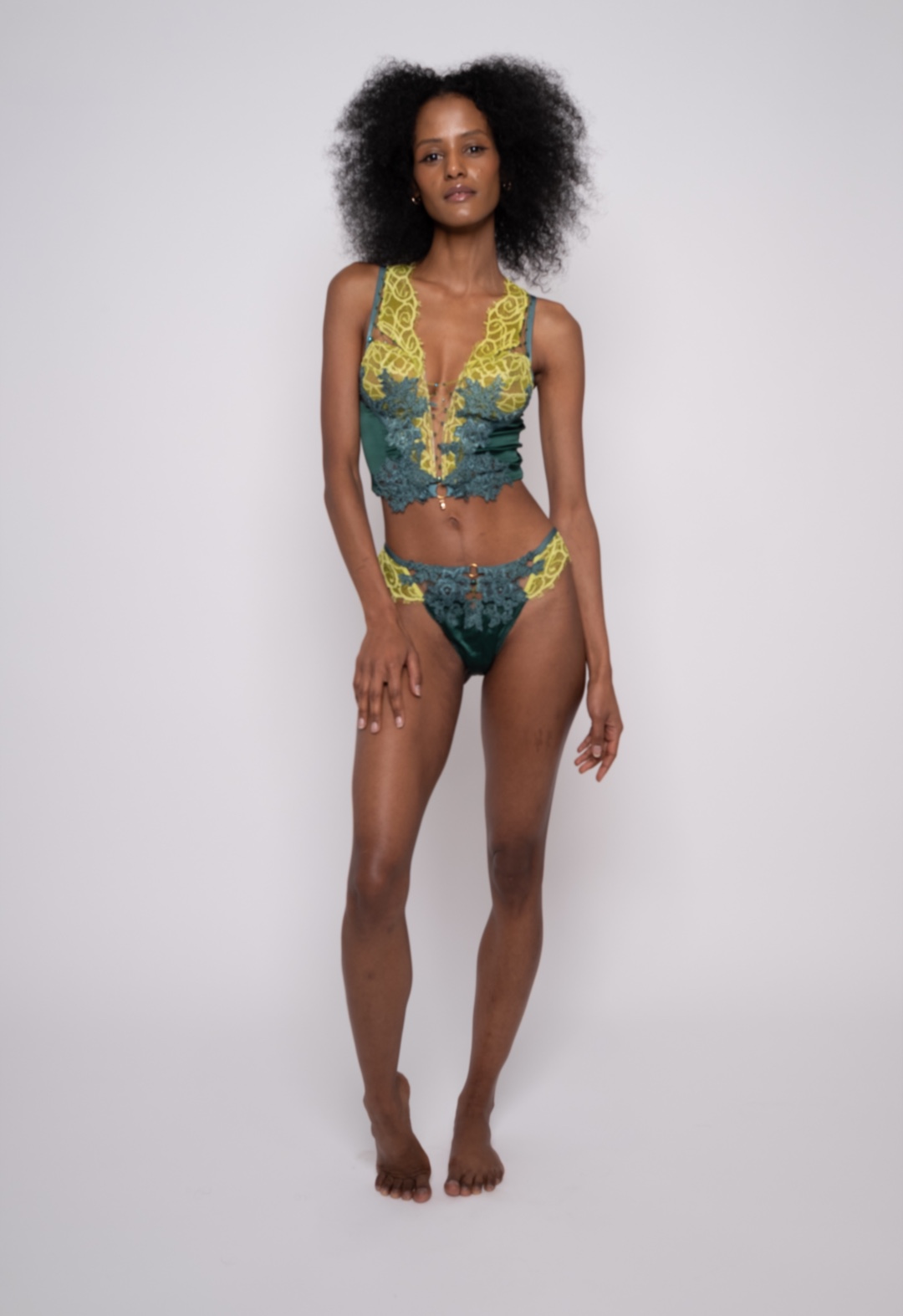 A slender black model facing the camera with one hip cocked, wearing an emerald and chartreuse bra and panty.