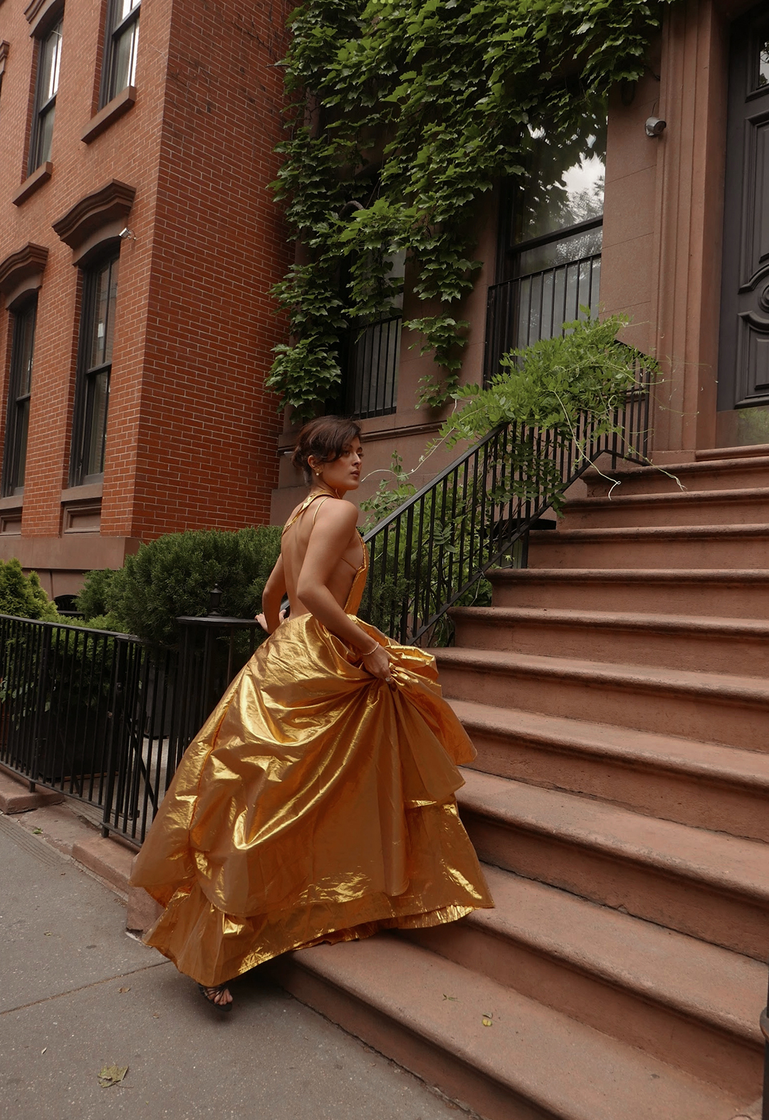 Photo of a model walking up the stairs of a Manhattan townhouse. It is a 3/4 back shot of her wearing an old Lamé one shoulder pleated a-line dress. The dress is backless. 