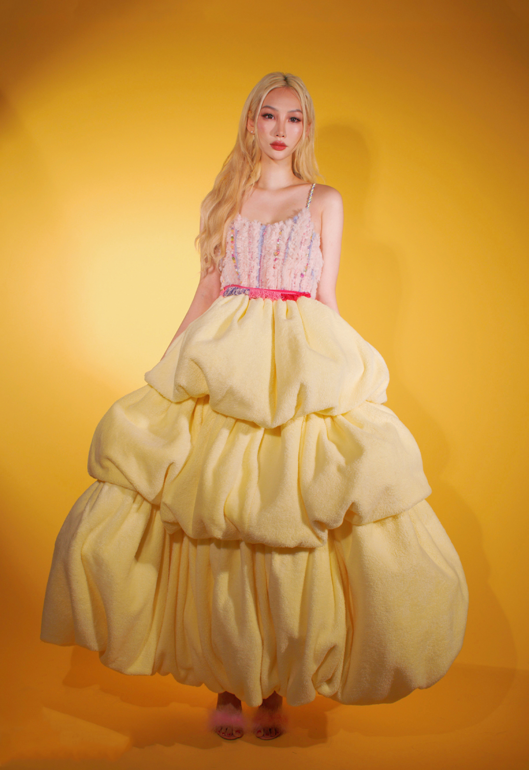 Photo of a girl with blonde hair standing facing to the camera. She has both of her hands holding the skirt of the dress. The dress she is wearing is a pastel pink-and-yellow furry dress. It has a bright-pink knit tassel belt. The background of this photo is yellow. 