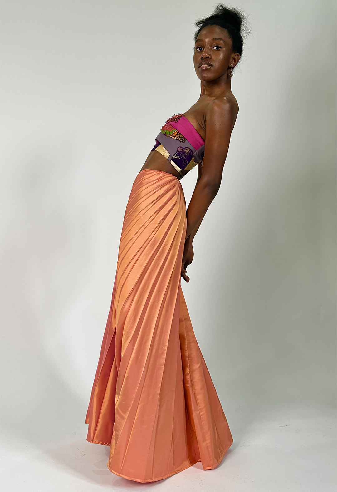 The side sunburst pleats cascade around the hips, fanning out and giving defined line direction, while model stands to the side, slightly leaning back with arms crossed behind their back.