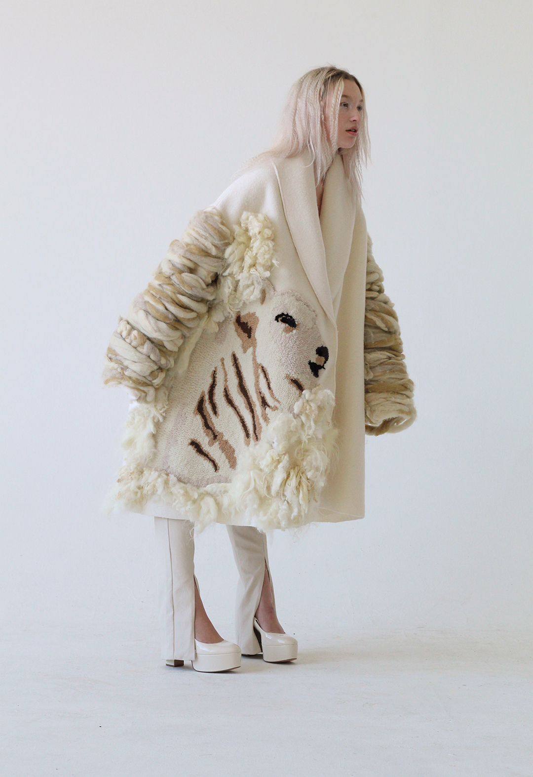 The model is standing and leaning forward, wearing a cream shawl collar coat with oversized drape, and tight, cream-colored stretch leather pants, rug-tufted sheep profile/portrait/motif.
