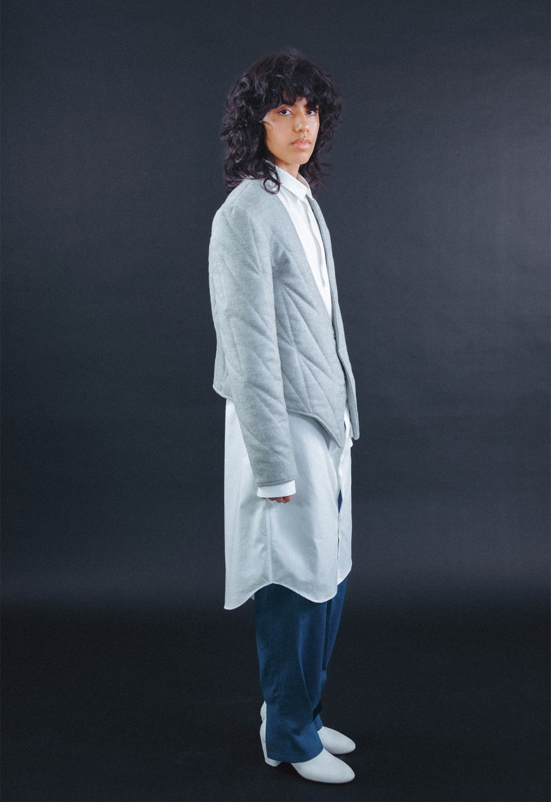 Photo of the model, Aria Puga, wearing a grey wool quilted jacket, a white cotton tunic shirt, and teal linen trousers. The model is turned to the side with their head turned towards the camera. The background is black.