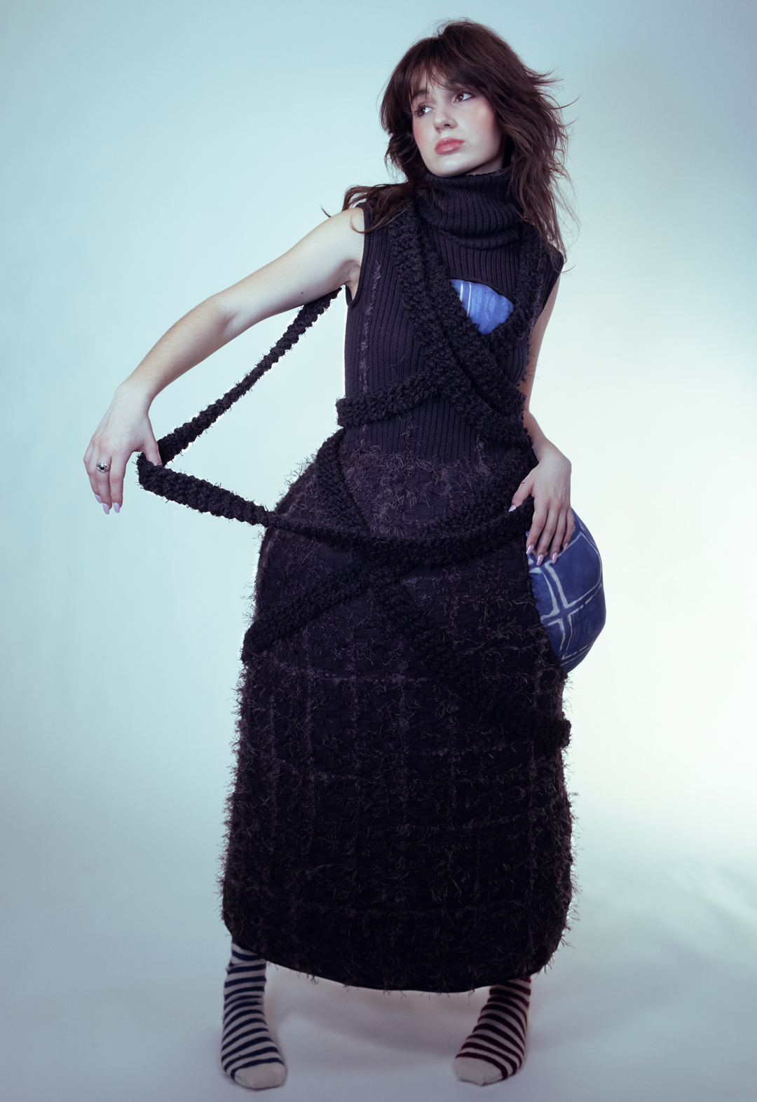 The model stands holding a long tassel out. She is wearing a black wide-hipped dress with textured embroidery on the bottom, along with blue-printed stuffed bulges on the chest and on the wearer's left hip. 