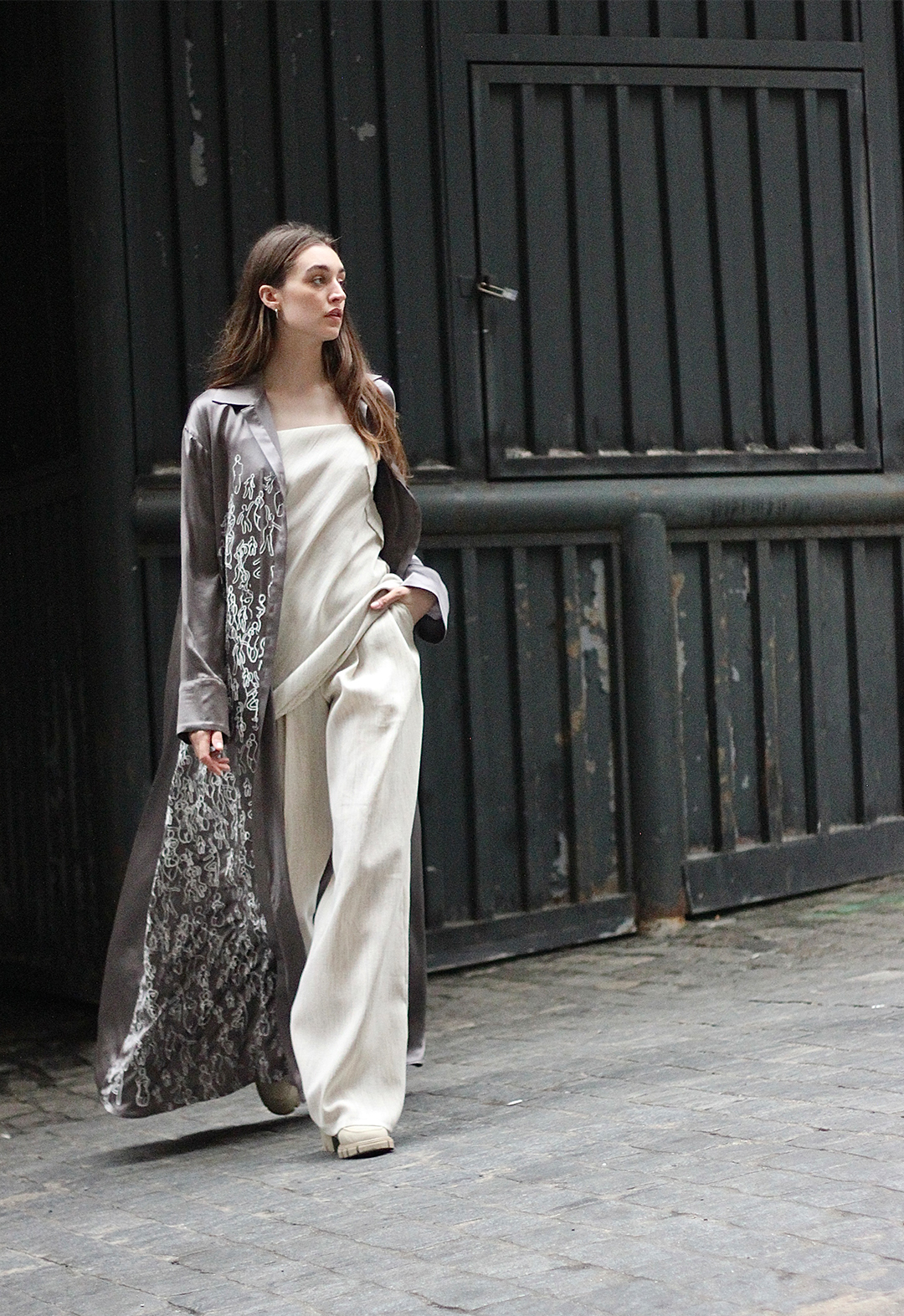 A model is walking while looking to the side with one hand in her pants pocket. She is wearing a gray printed silk shirtdress over a cream slip dress and matching wide-legged pants.