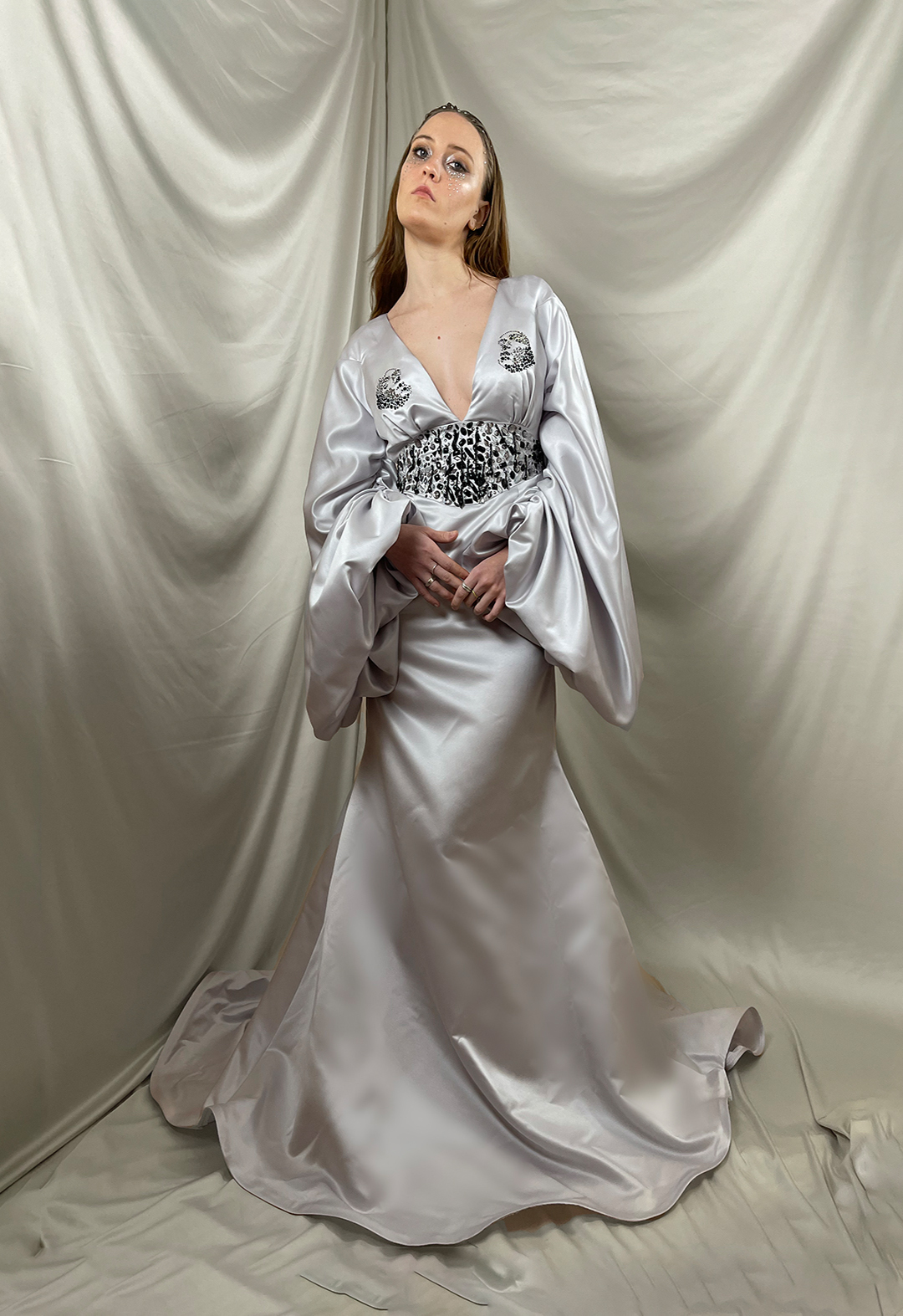 A model wearing a silver mermaid gown with beaded yoke, a deep V-neckline and a drop-shoulder bishop sleeves with gathering. The background has champagne color silk drapes.