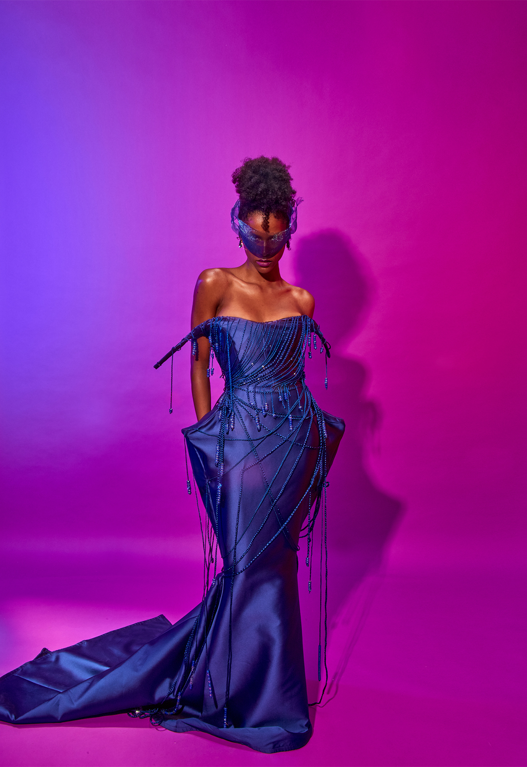 Blue purple background with the model standing alone. She is wearing a blue sculpted gown. 