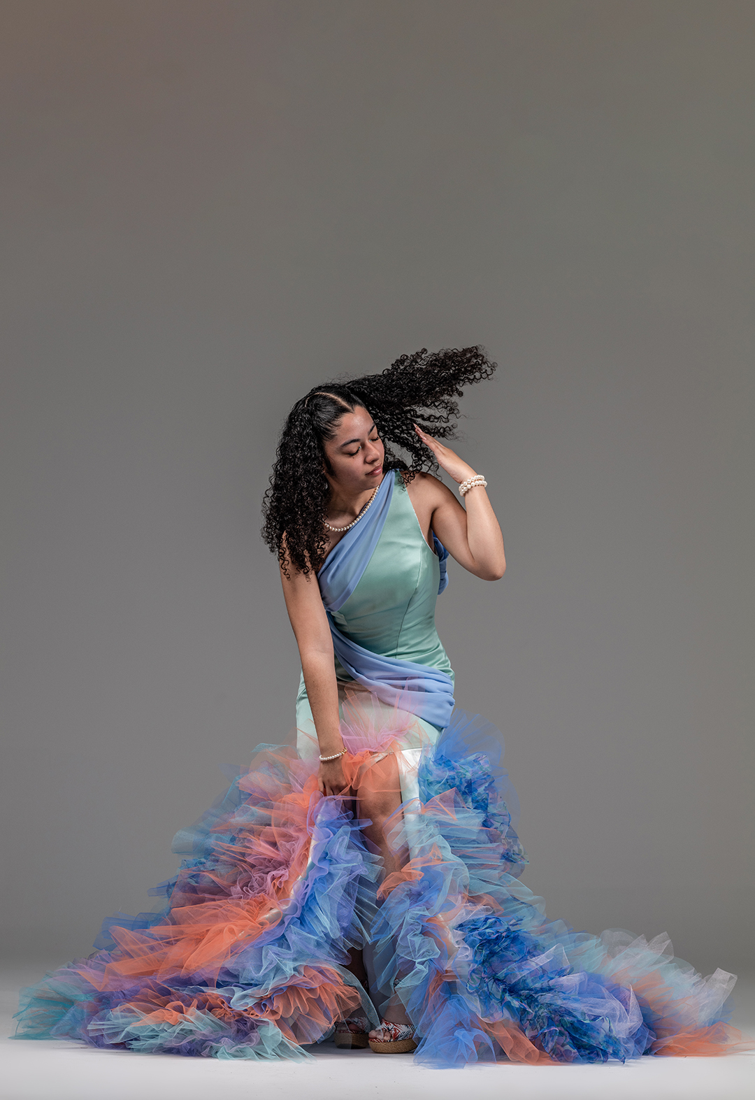 Front view of a model bending forward, wearing an asymmetric mermaid evening gown with tulle ruffles and a slit on the bottom. The model is flicking her hair back. The background is gray. 