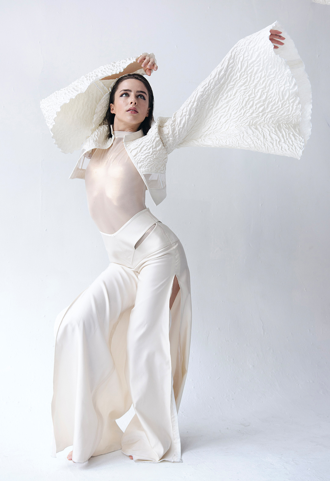 This is a photo of a model wearing a seashell white matelassé organza kimono sleeve jacket, and cut-out silk satin flared pants. She has both of her hands diagonally up towards the right side of the photo. Her knees are bent and her hip is out towards the right. 