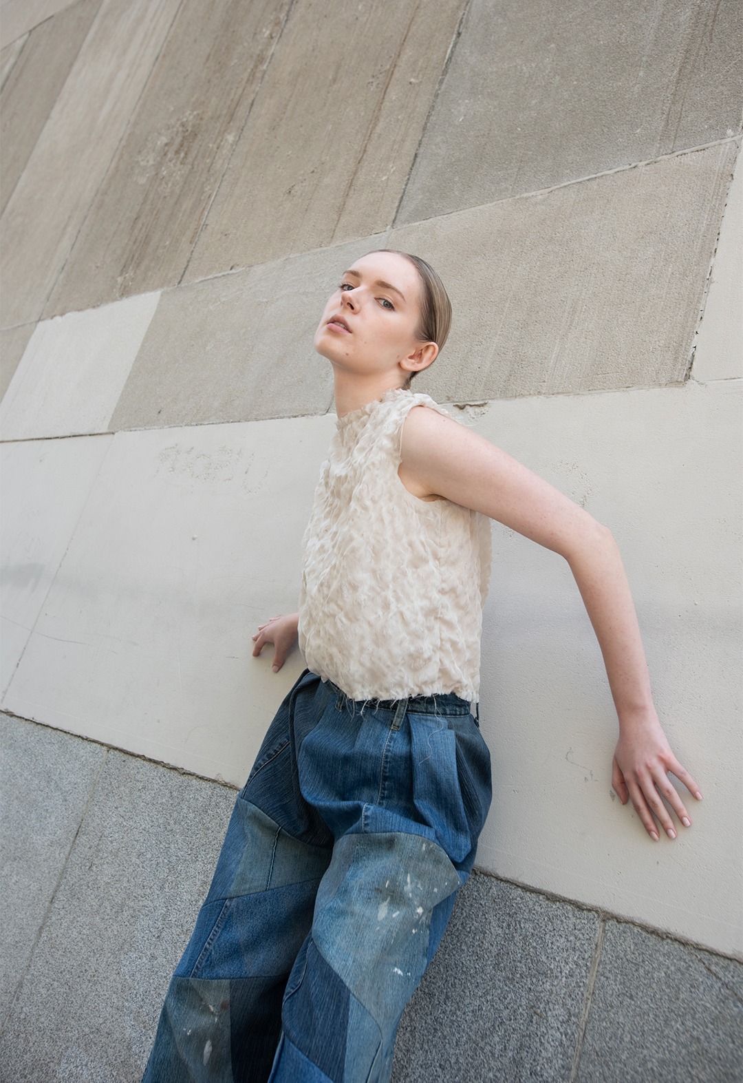 This blouse is made by hand manipulating and stretching apart the georgette silk fabric. The photo is of a girl wearing a beige distressed blouse with patchwork upcycled denim blue pants; Her torso is at an angle, slightly turned from the front and leaning against a concrete wall. The background has multicolored gray and beige concrete panels. 