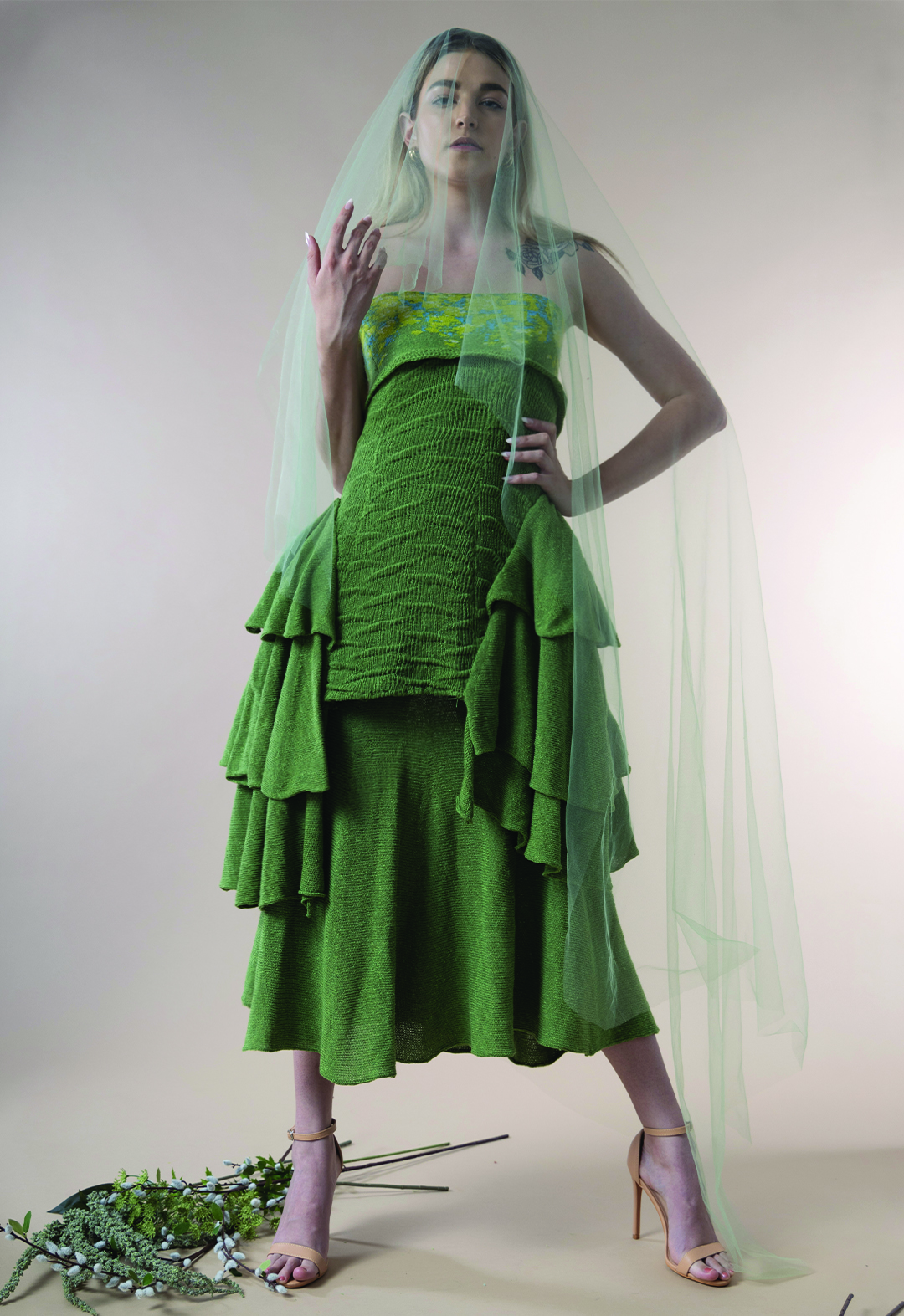A female model is posed in a front view in a midtone green gown with a tube top, bumpy textured bodice, and layers of flare skirts attached in different lengths on each side.