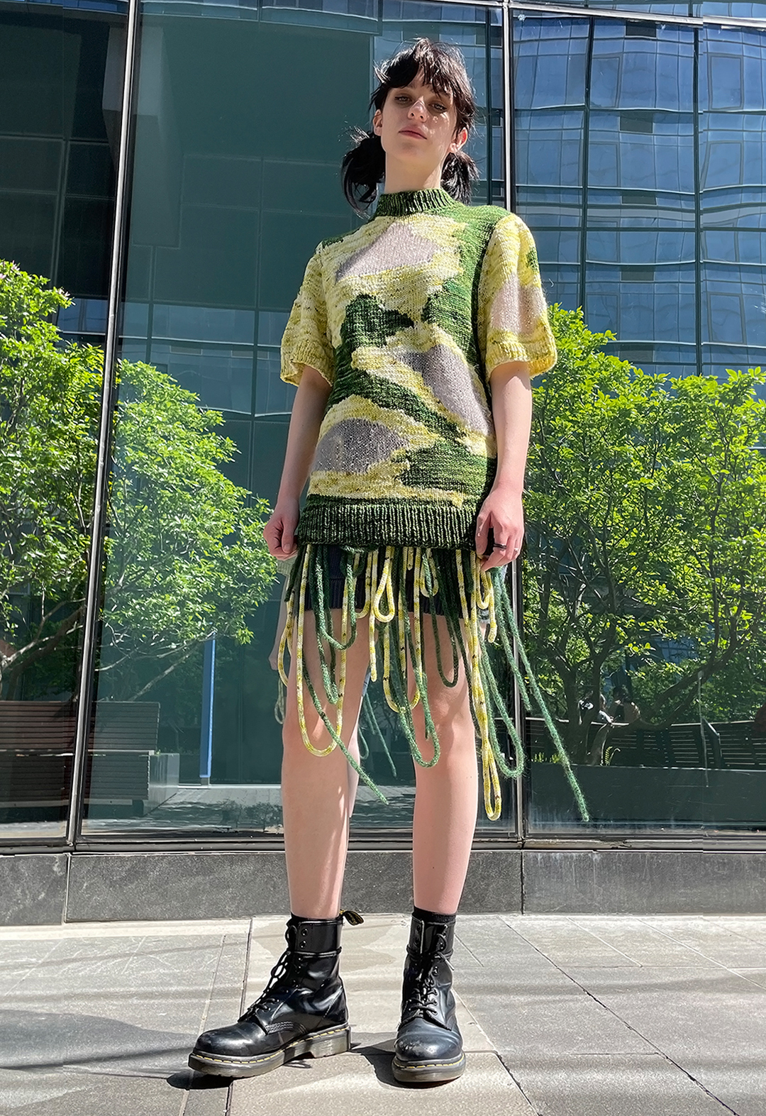 Front view of a model wearing a hand knit short sleeved intarsia sweater with transparent mohair windows. She is also wearing combat boots. There is a glass building in the background with reflection of trees.