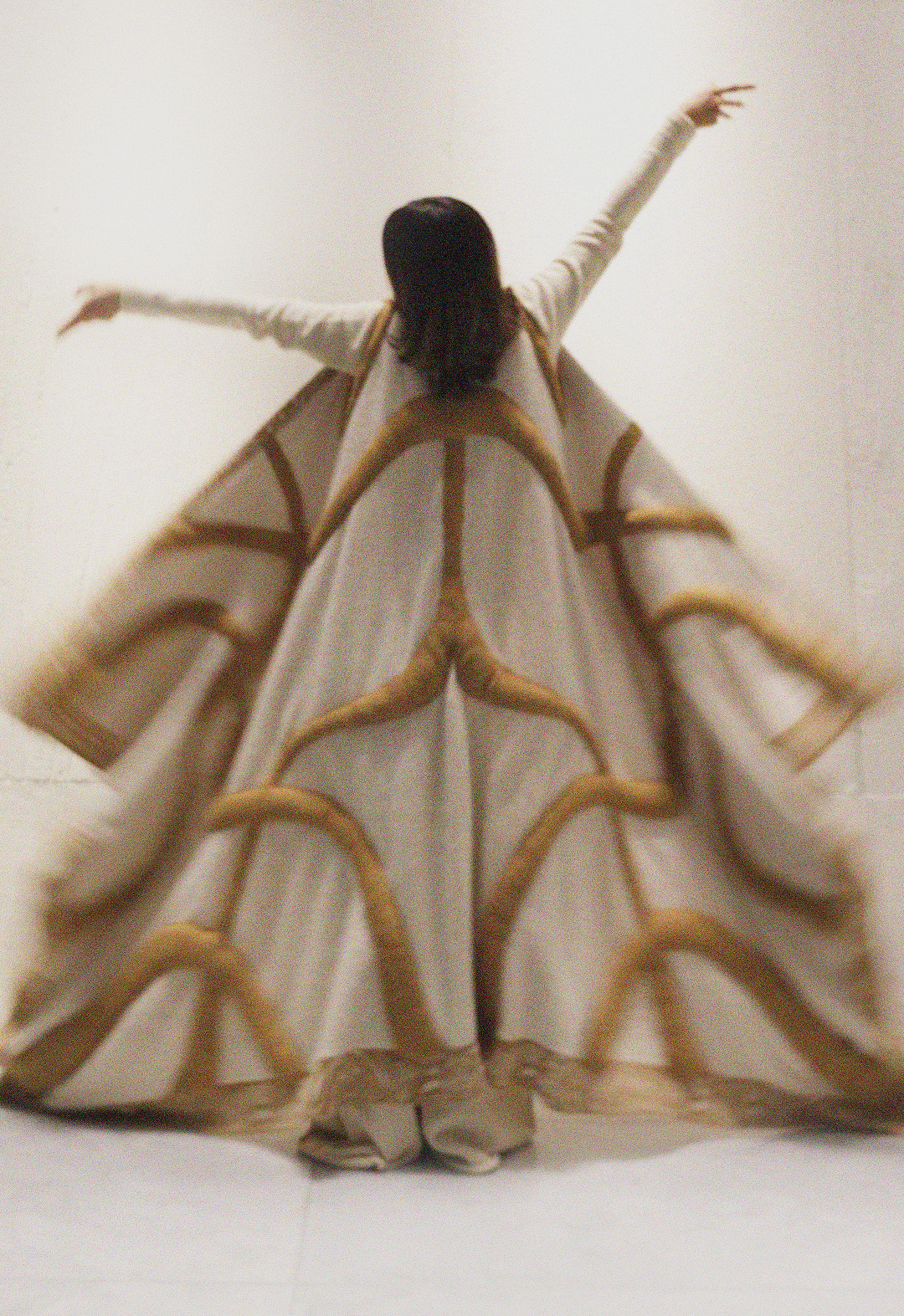 The model is wearing a huge two-color, jacquard pattern reversible cardigan, which has borders all around. The tan pattern lines inspired by sari are stuffed. She is wearing white wide-legged pants, which are a little bit visible below the hemline of the cape. Her hands are up in the air, and the background is white. 
