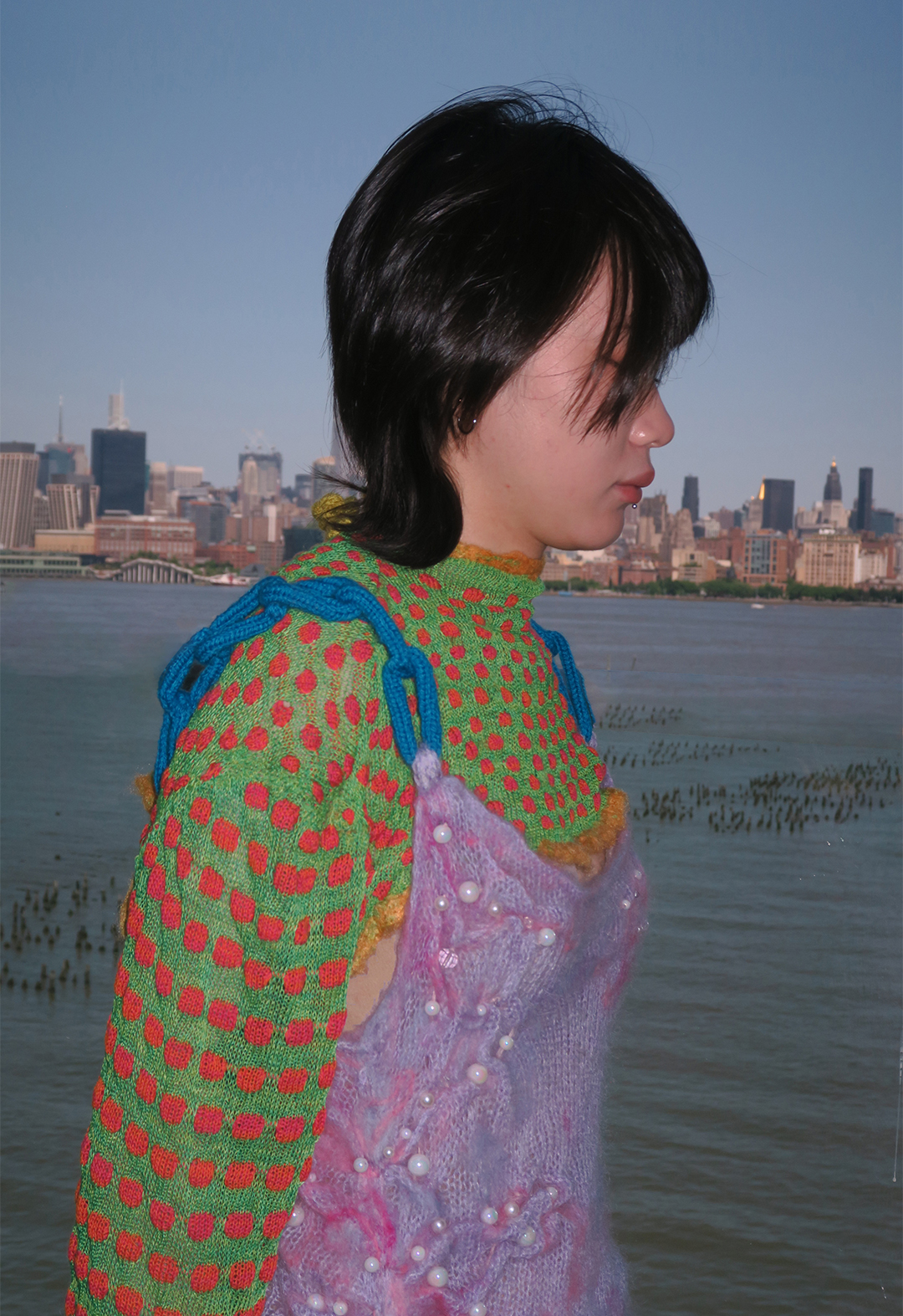 Close up side view of a model wearing a green sweater with red polka dots and a pink spaghetti straps knit dress over the sweater. The Manhattan skyline is in the background. 