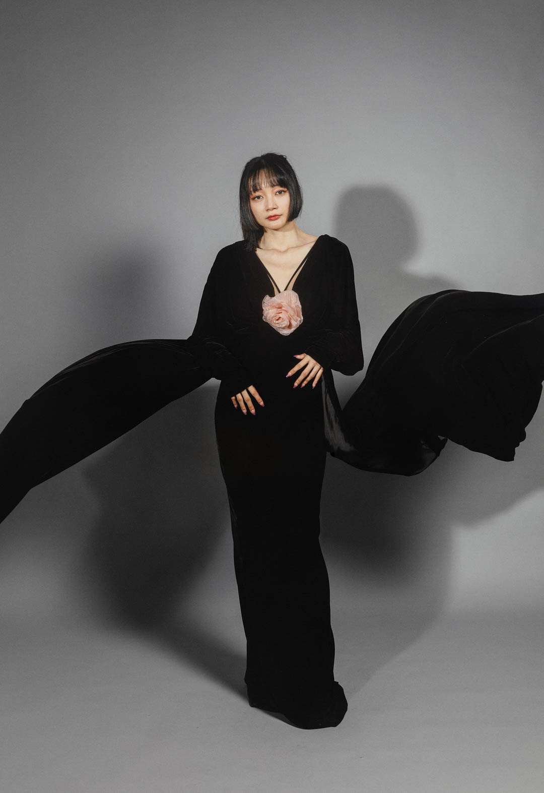This black velvet dress features a V-shaped neckline and narrow straps on each shoulder for support. The dress is long, with a flowing train that drapes between the wearer's legs. The background in the image is gray.