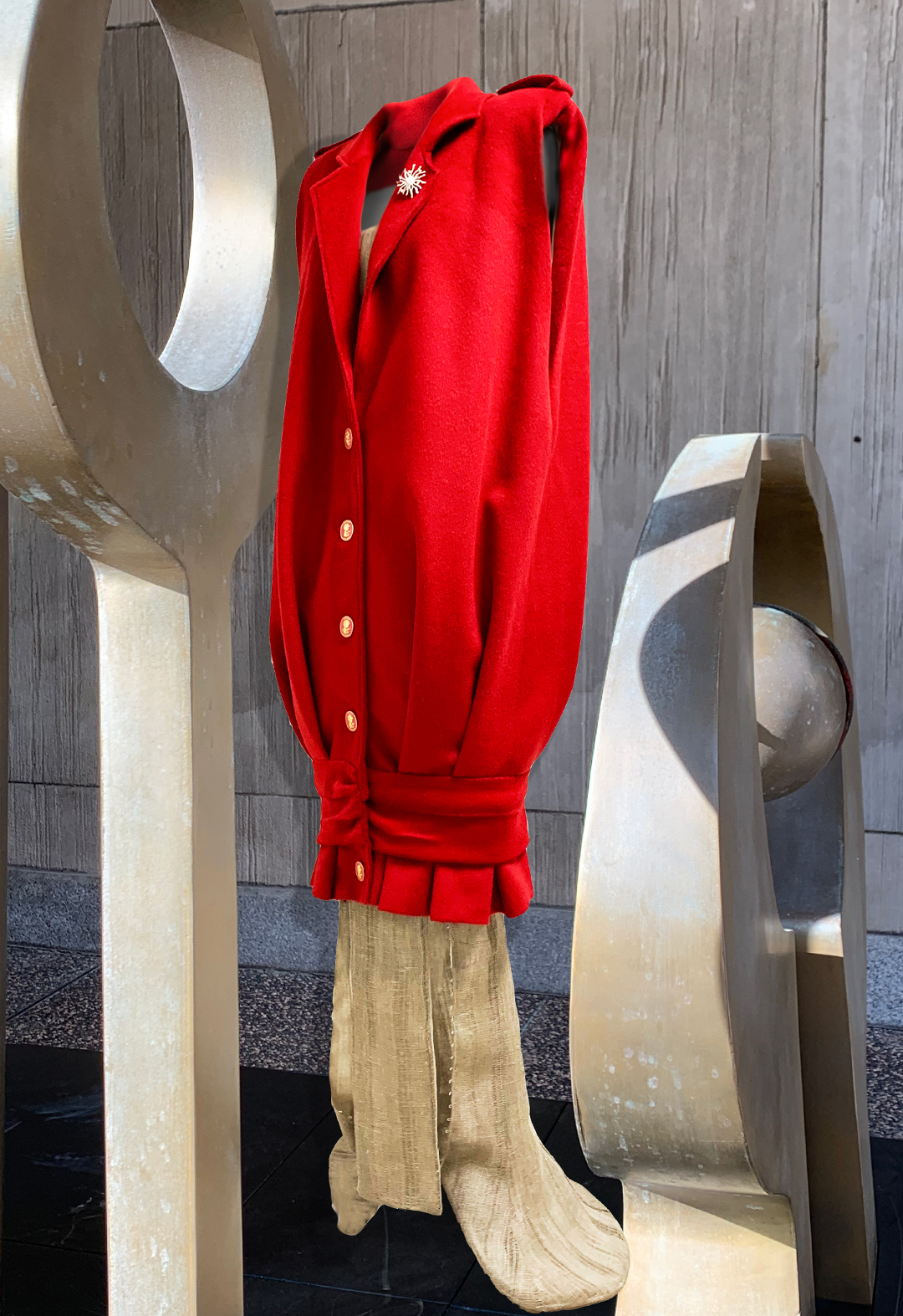 The top is a long red vest. There is a linen dress underneath. The clothes are presented against a background of a group of metal sculptures.