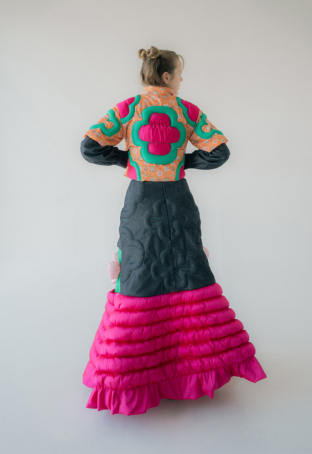 This image shows the back view of the short puffer jacket and flared skirt. The jacket is quilted in floral motifs that are reddish-pink and bluish-green color. The denim skirt is mixed and matched with taffeta fabric that was divided into six lines and filled with polyfill.