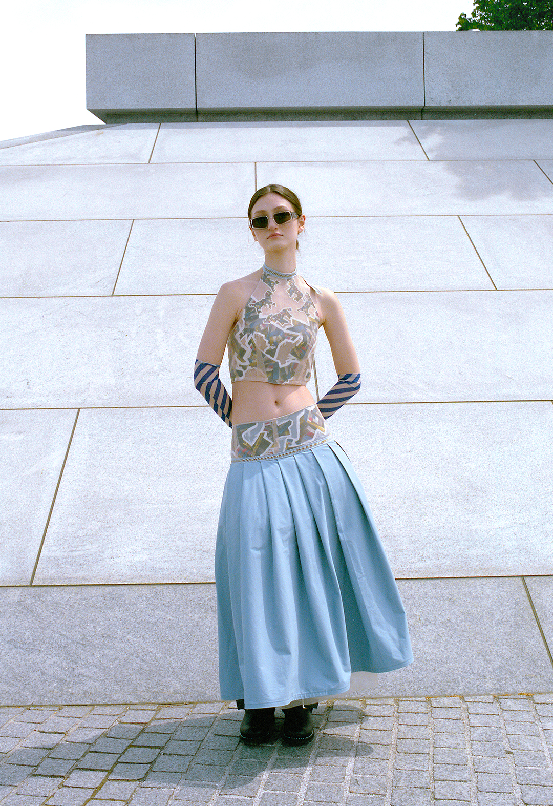 This two-piece set includes a halter neck crop top and knife pleat yoke skirt. The top and yoke parts are appliquéd with a patchwork digital print stitched between organza and mesh. Lacing detail on the back for opening. Accessorized with dark-blue mesh long-sleeved gloves and sunglasses. The model is posed in front of a gray wall.