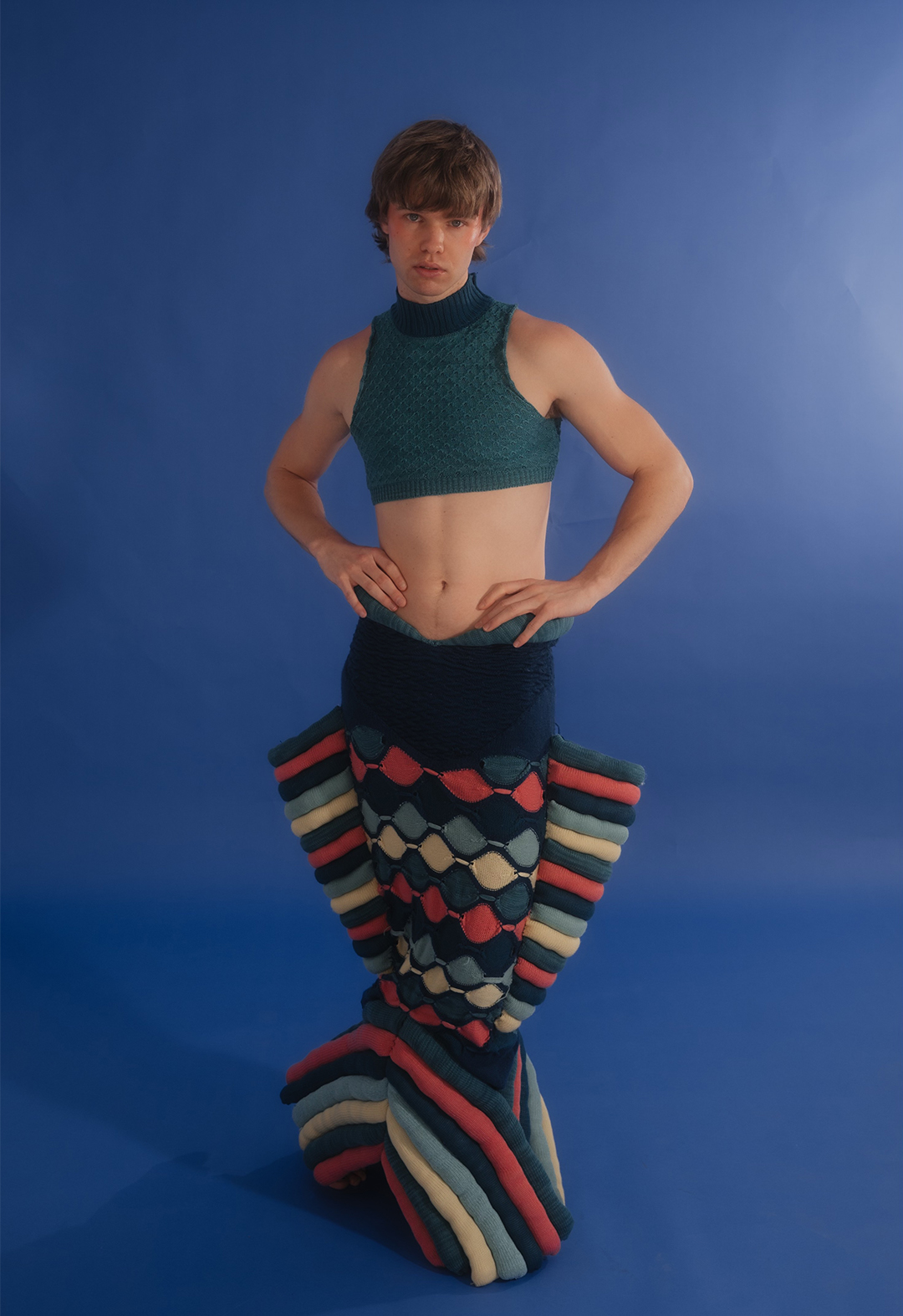The model is facing front and wearing a blue, multicolored sculptural fish skirt and teal knit tank top.