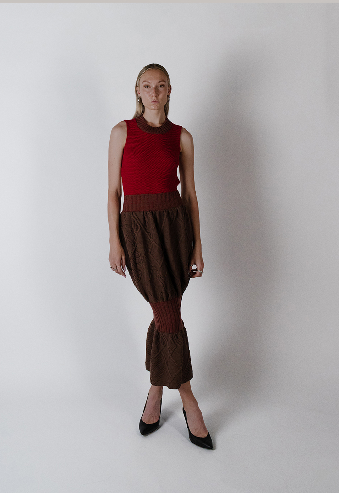 This is a red-and-brown sleeveless mock neck dress. There are two heather red and brown rib sections, at the waist and the knee, creating a balloon effect on the skirt. The top of the skirt is bright red with moss stitching. The balloon skirt is filled with irregular Aran cables, with a mix of moss, link links, and ribbing. The model is standing in black high heels with her legs crossed. The background is white.