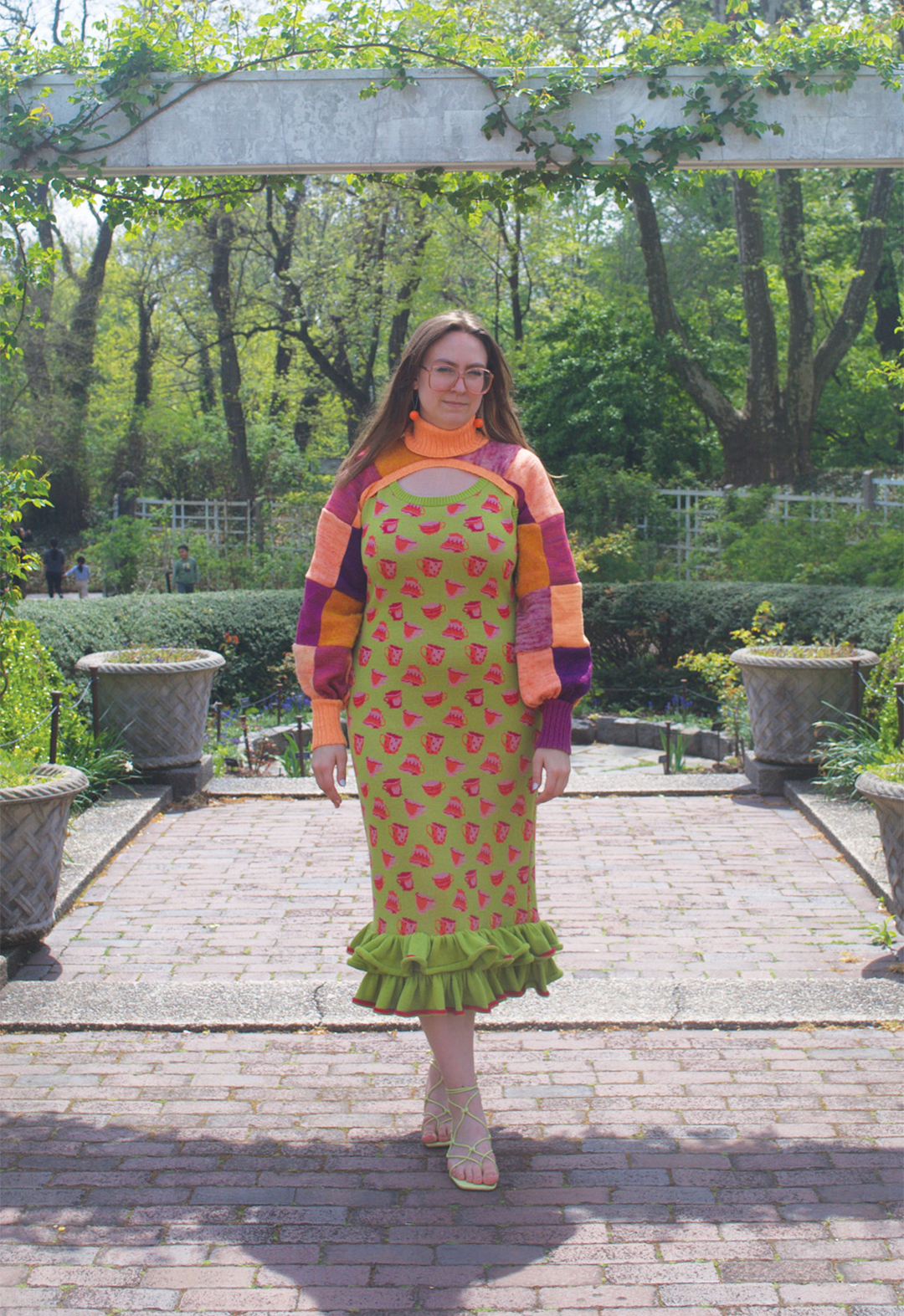 Photo of a model wearing a green, teacup-motif dress with a ruffle hem and a purple-and-orange patchwork turtleneck shrug and standing under an archway.