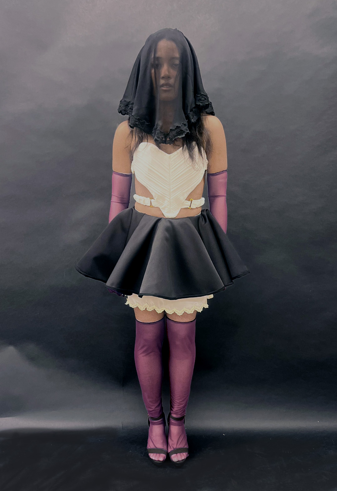 Full-body shot of a model staring straight at the camera. She wears a black draped, embroidered veil, an ivory heart-shaped bustier, a two-tiered black-and-white miniskirt, deep-pink gloves, and thigh-high socks.