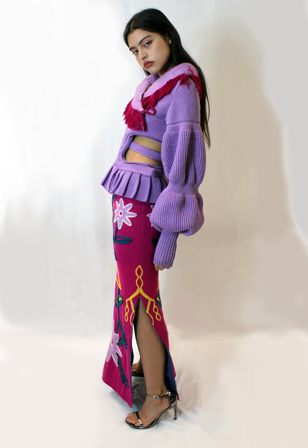A model standing against a white background. She has long hair and is wearing a lavender color knit wrap top with shaped sleeves and crochet fringe at the shawl collar. She is also wearing a skirt with a floral jacquard multicolor pattern.