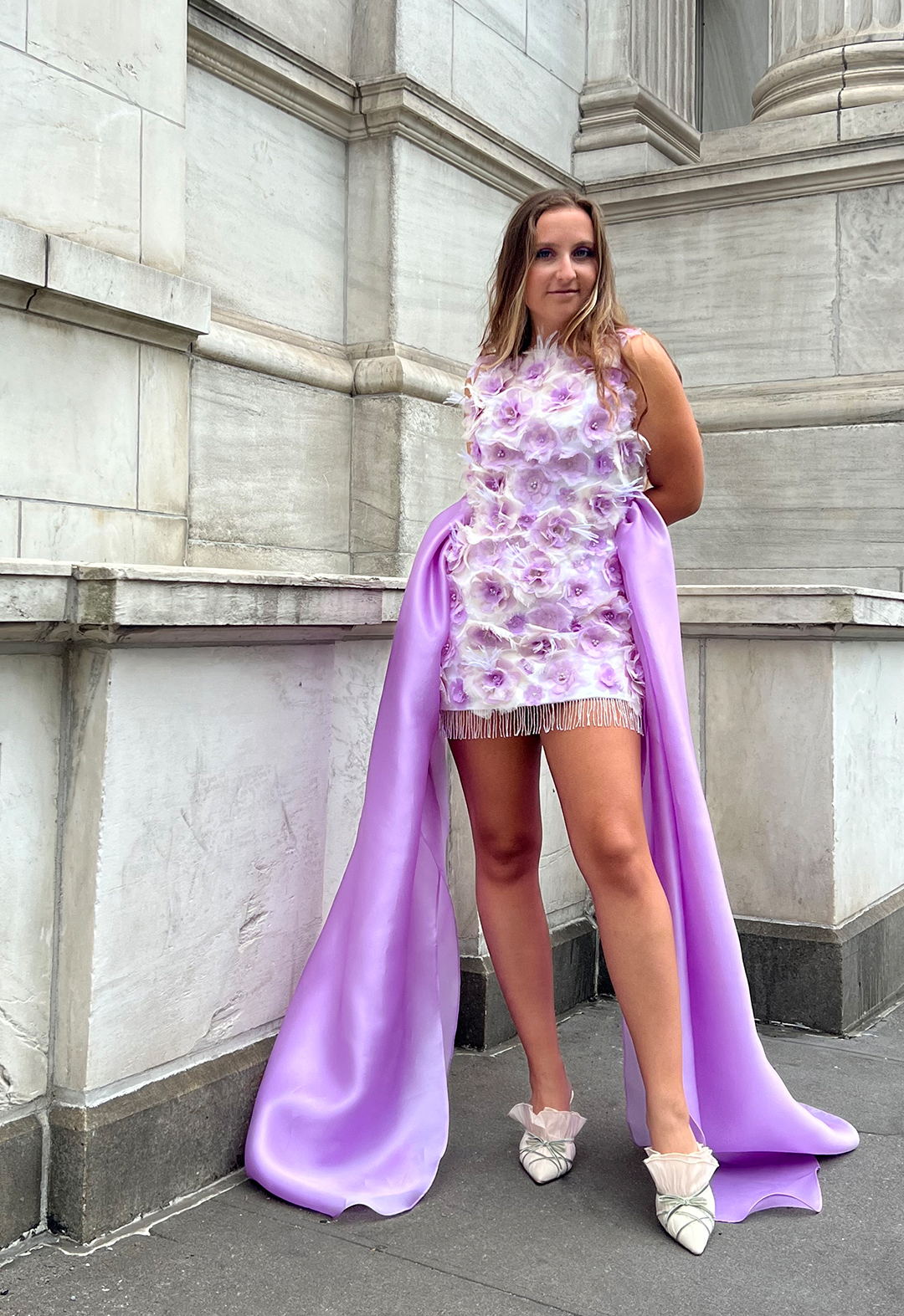 This dress is made of silk satin, organza, and chiffon. The photo shows a girl wearing a white satin mini dress with lilac embellished chiffon and organza flowers and a beaded fringe hem. She is smiling and her hands are behind her back.  Gathered lilac organza extends from the side seams to the ground. The background is a white marble building. 
