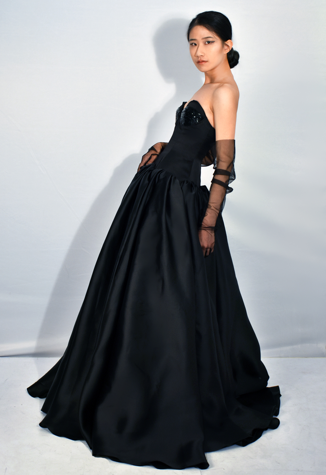 This black gown is made from 100 percent silk. The photo shows a side view of a woman wearing a black silk gown with beading covering three-quarters of the bodice cups. The floor-length gown has a drop-waist duchesse satin bodice with cone-like satin-faced organza skirt panels gathered along the drop-waist, pooling onto the floor. The model is wearing black tulle gloves that cover three-quarters of her arms. Her face is facing the viewer. The background is white.