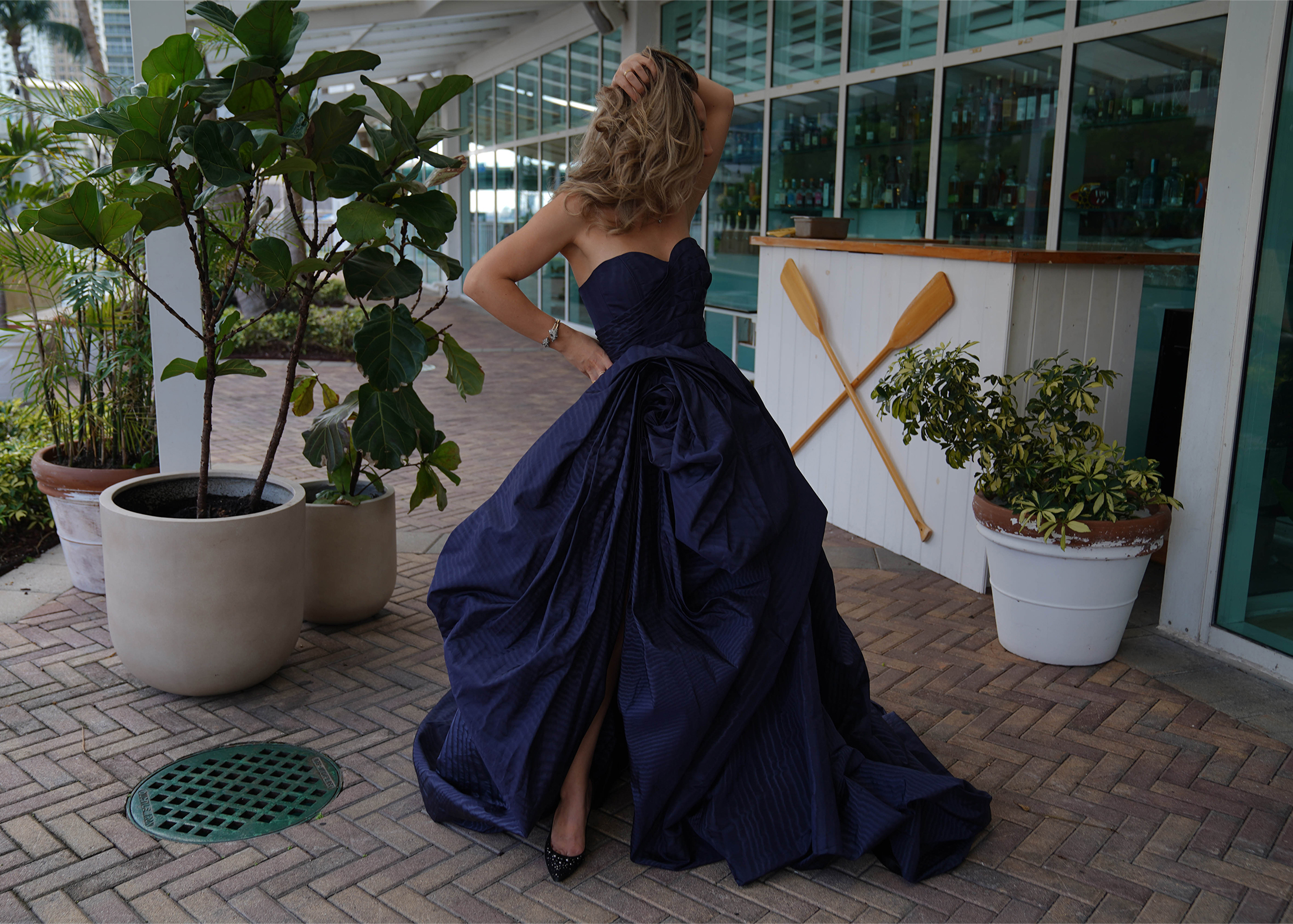 A model is standing around potted plants and looking towards her right. She is wearing a dark blue moire faille cowl skirt and fluted bodice gown.