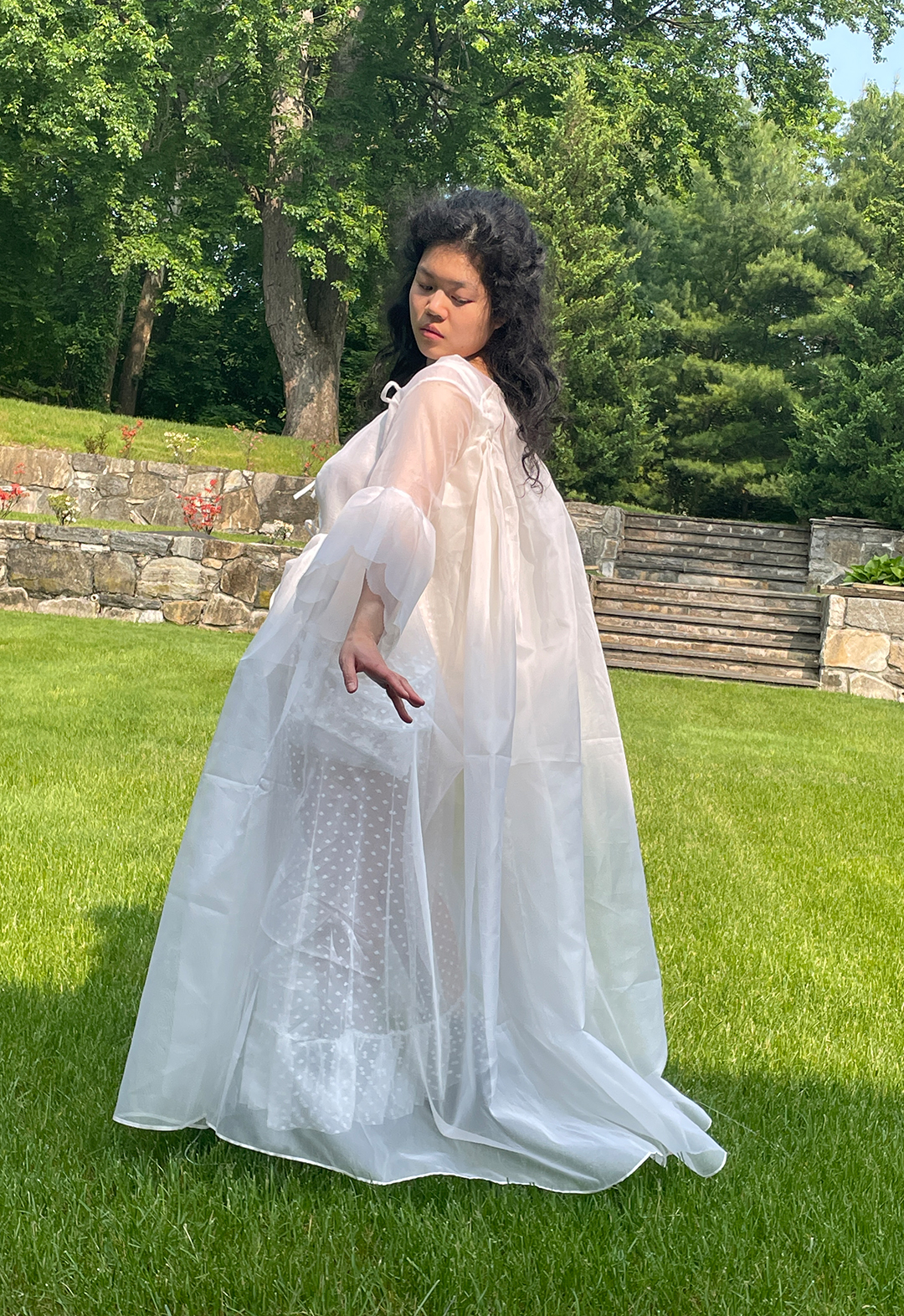 Side view of a model standing in a garden and wearing a white organza robe with puffed sleeves.