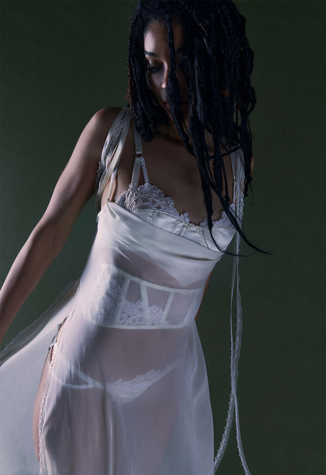 This image features an up-close view of a model in a long pearl-white bias gown with a cowl neck, open side seams and lace detailing. It is paired with a bra and panty set and matching waist corset embellished with lace and pearls. The image is set in a green background.