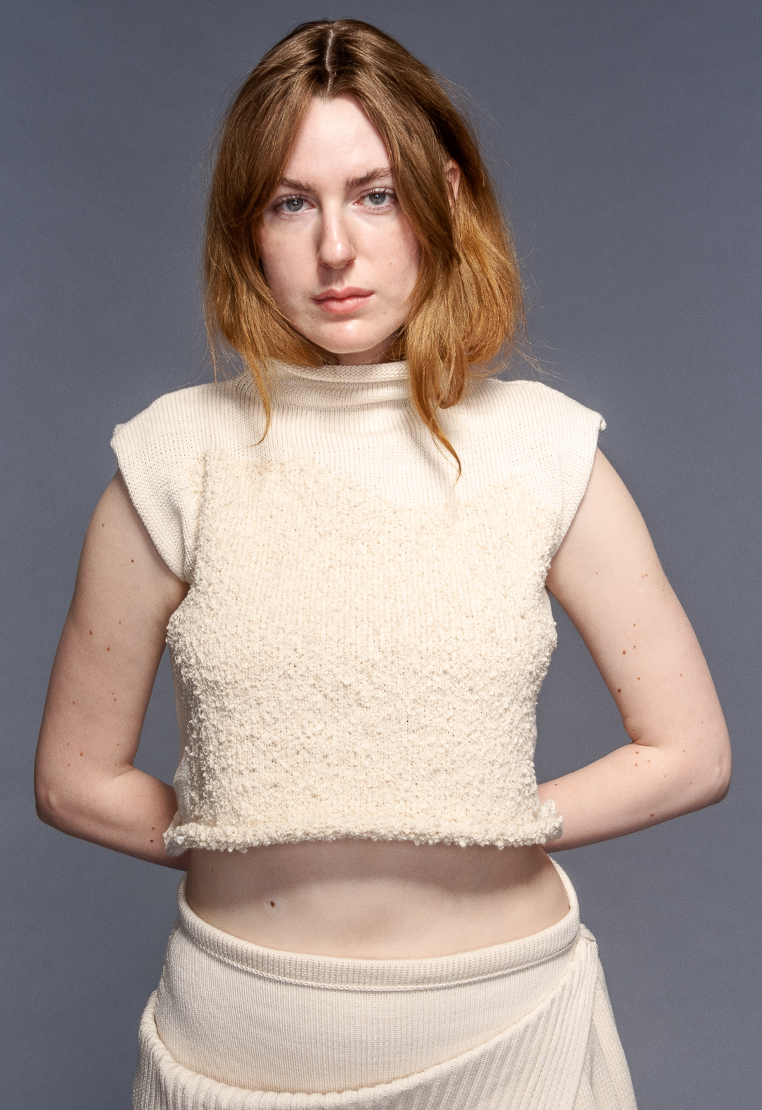 Model wears funnel neck top with misshaped intarsia and irregular pleated skirt in organic cotton. Her hair is tucked in the sweater.