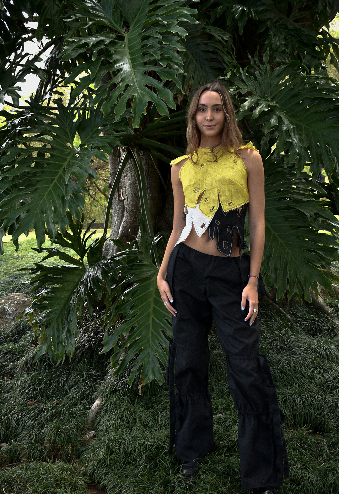 Yellow/black/white "monstera"-shaped cut-out burlap top, paired with canvas low-waisted drawstring pants. Model is standing in front of a big tree full of monstera leaves.