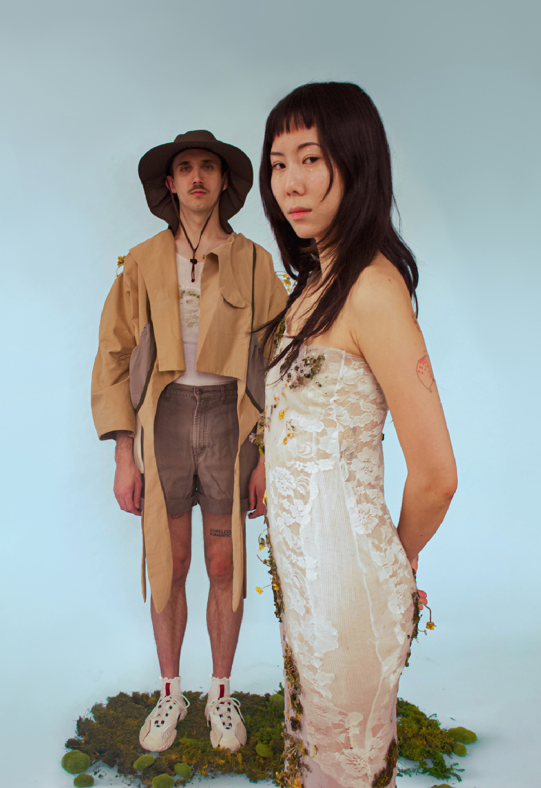 In this photo, the female model stands close to the camera, while the male model stands far behind. Although both models are looking at the camera, the female model has her body turned to the side while keeping her eyes on the camera.