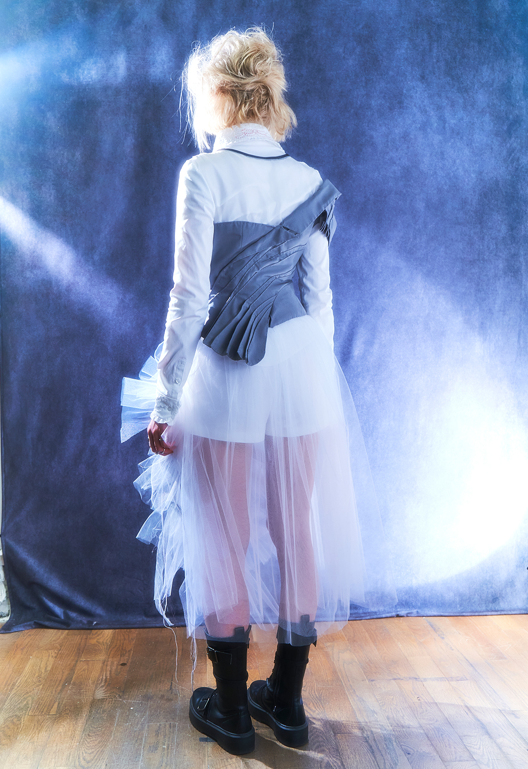 Back view of the design. The blonde model stands in front of a blue backdrop in a sheer dress worn over a blouse and short with an attached jacket.