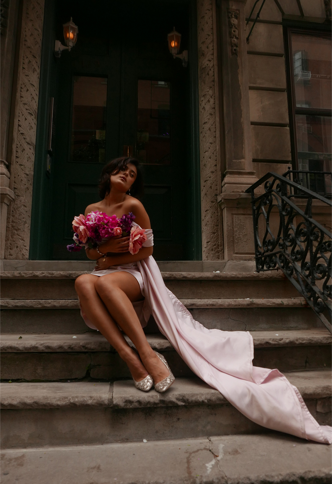 Photo of a model sitting on the stairs of a Manhattan townhouse. She wears a baby pink off-shoulder dress with draped cowl and side train with flowers on the neckline.