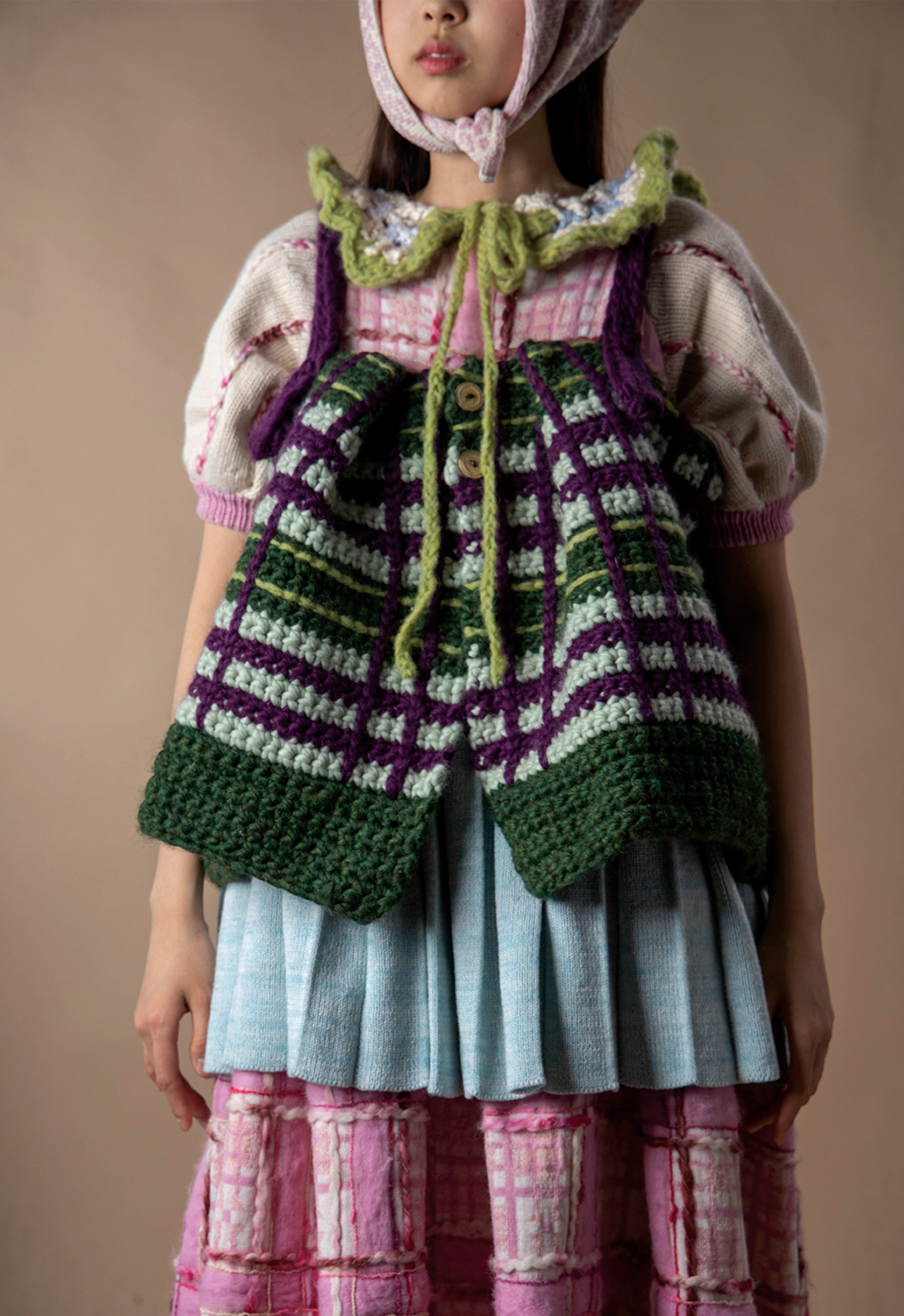 A close-up shot of the model wearing five layers of knitted garments: an apron top, a long dress, a pleated skirt, a collar, and a bandana on her head. 