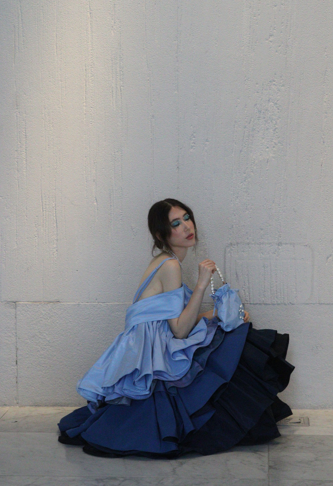 Model sitting on a marble floor, wearing an off-the-shoulder silk taffeta gown with ruffles in multiple shades of blue.