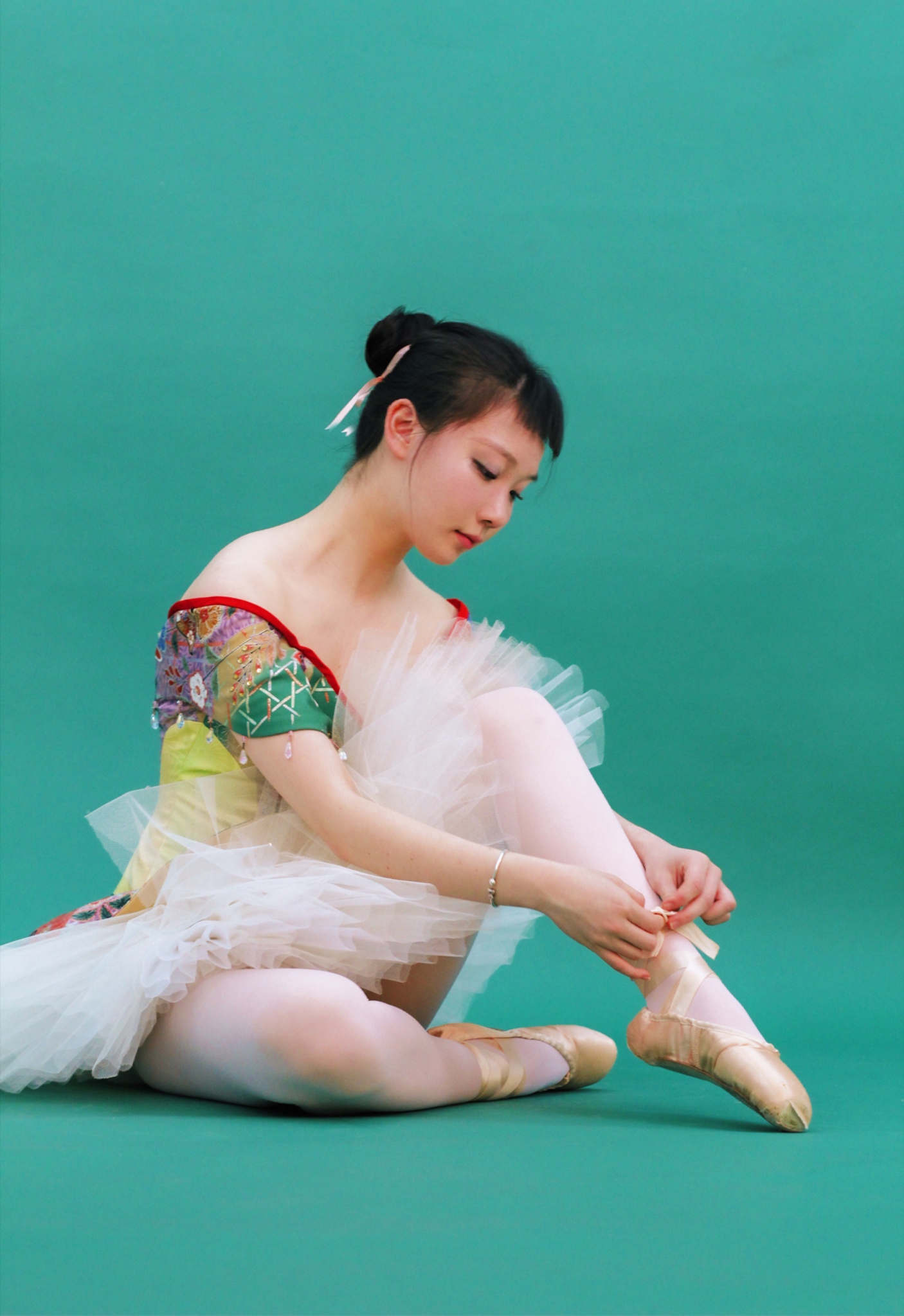 The photo shows a ballerina in a kimono-inspired ballet tutu sitting on the floor tying her pointe shoes. 