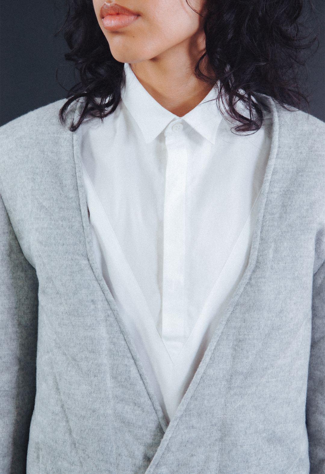 Front-close-up photo of the model, Aria Puga, wearing a grey wool quilted jacket and a white cotton tunic shirt. The neckline opening of the jacket corresponds with the geometric hidden half-placket opening of the shirt. The background is black.