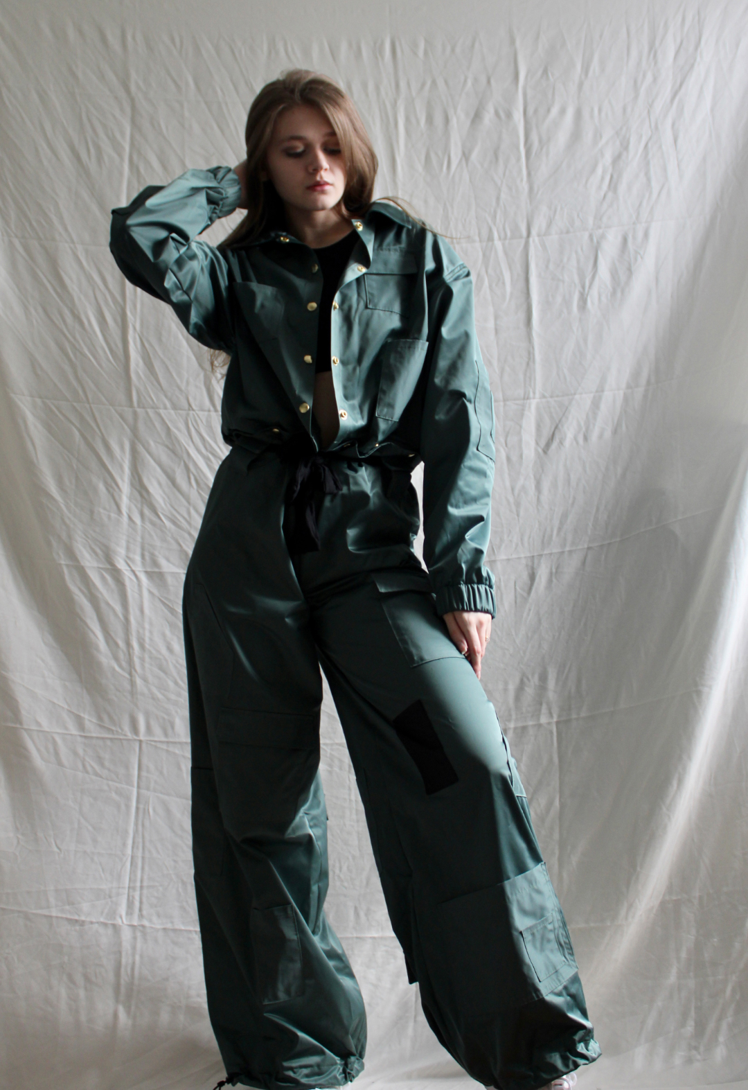 Front view of a model wearing a deep green, nylon jumpsuit that buttons at the waist. She has one hand on the side and the other hand bent up, behind her head. 