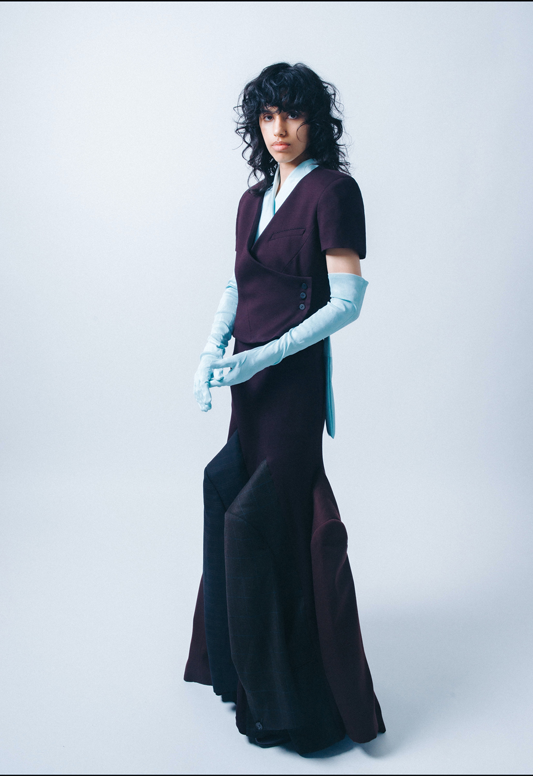 The photo presents a three-quarter view of Look 1, which consists of a dark-purple short-sleeve dress suit with wavy seams and re-engineered blazers, and features an exaggerated shoulder cape with gathered drape.